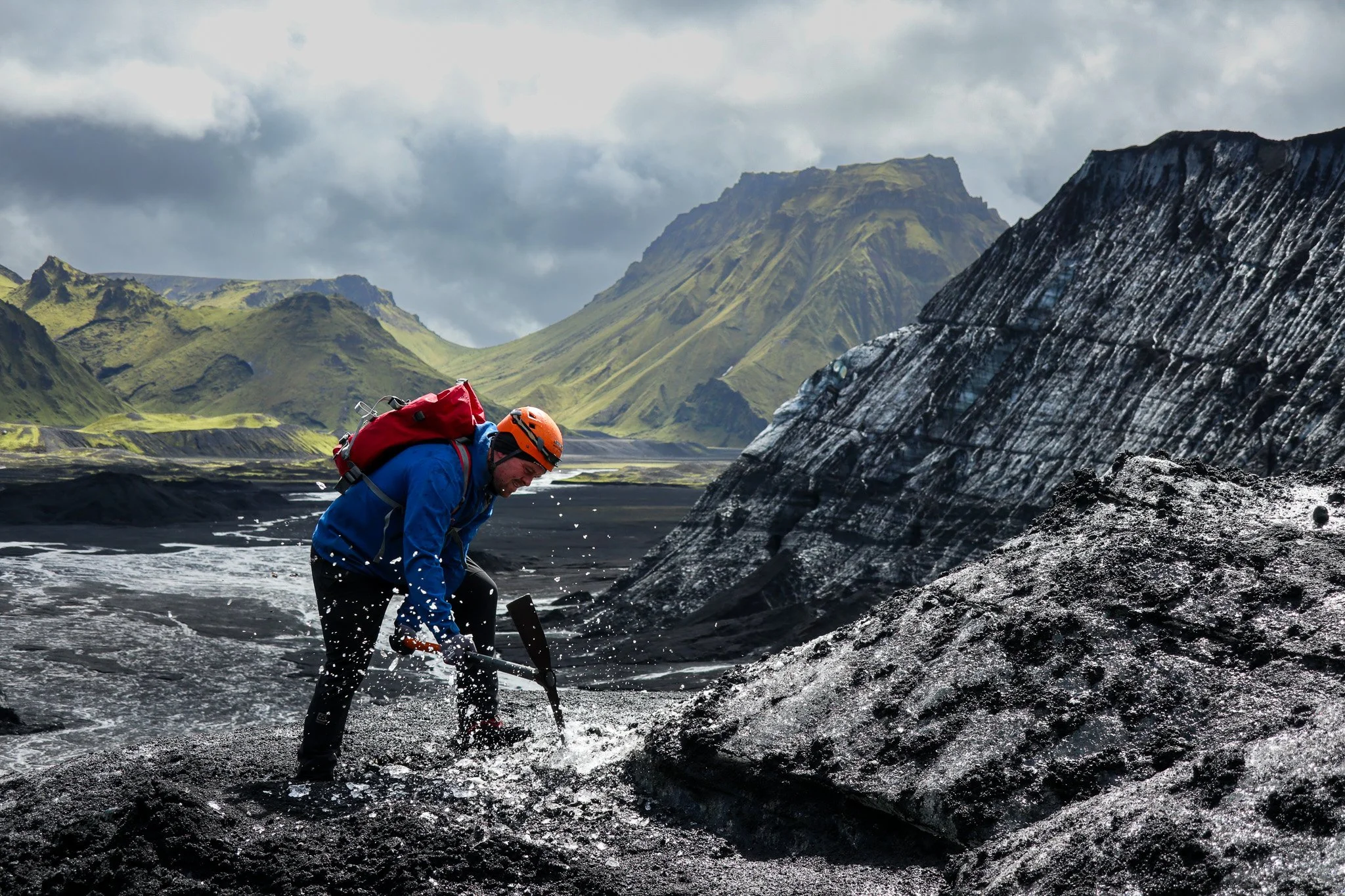 A person wearing a blue jacket, orange helmet, and red backpack digging in black volcanic sand with a pickaxe in a rugged volcanic landscape with green mountains in the background.