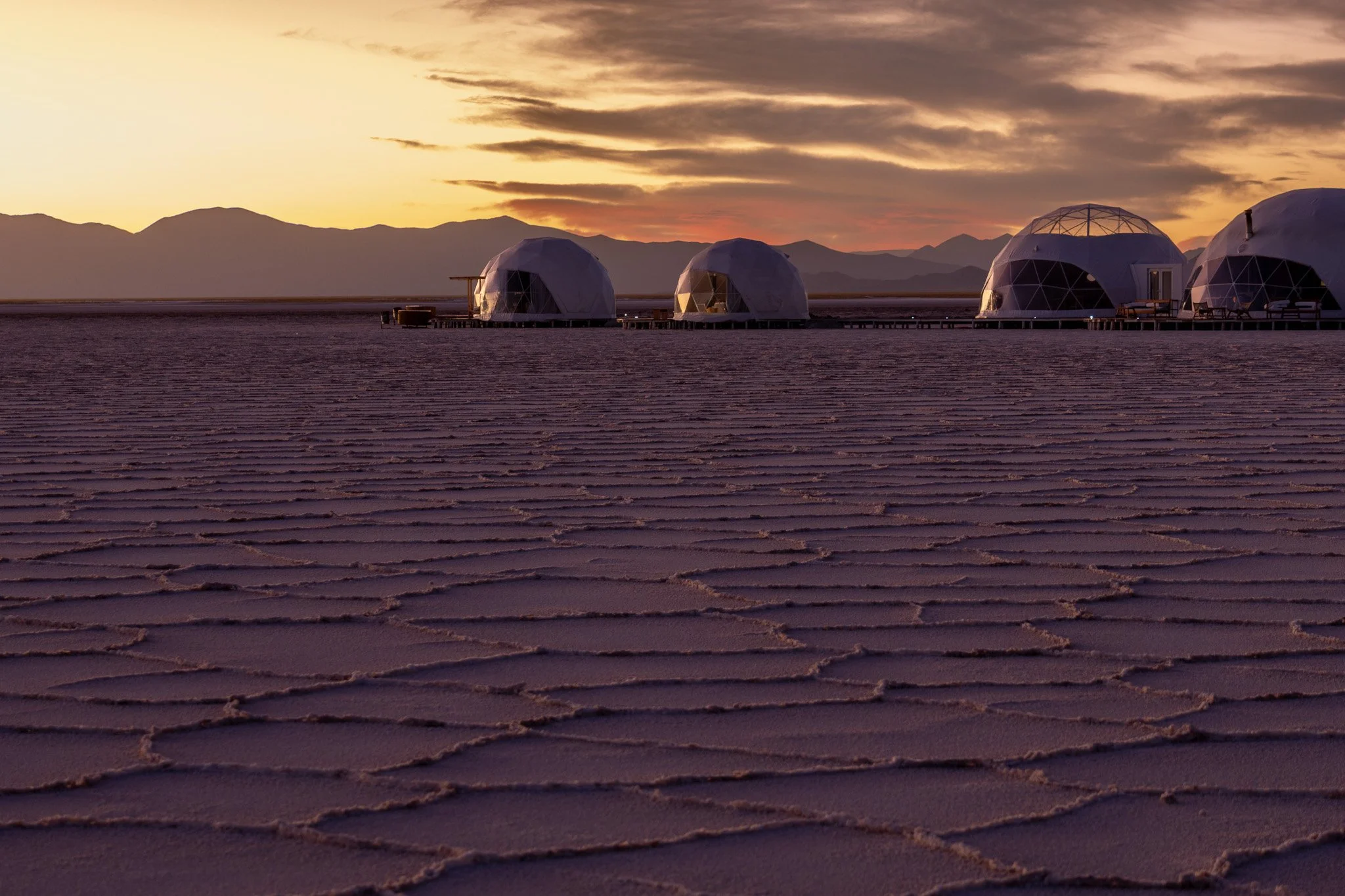 Four geodesic dome structures on a salt flat with mountains in the background during sunset.