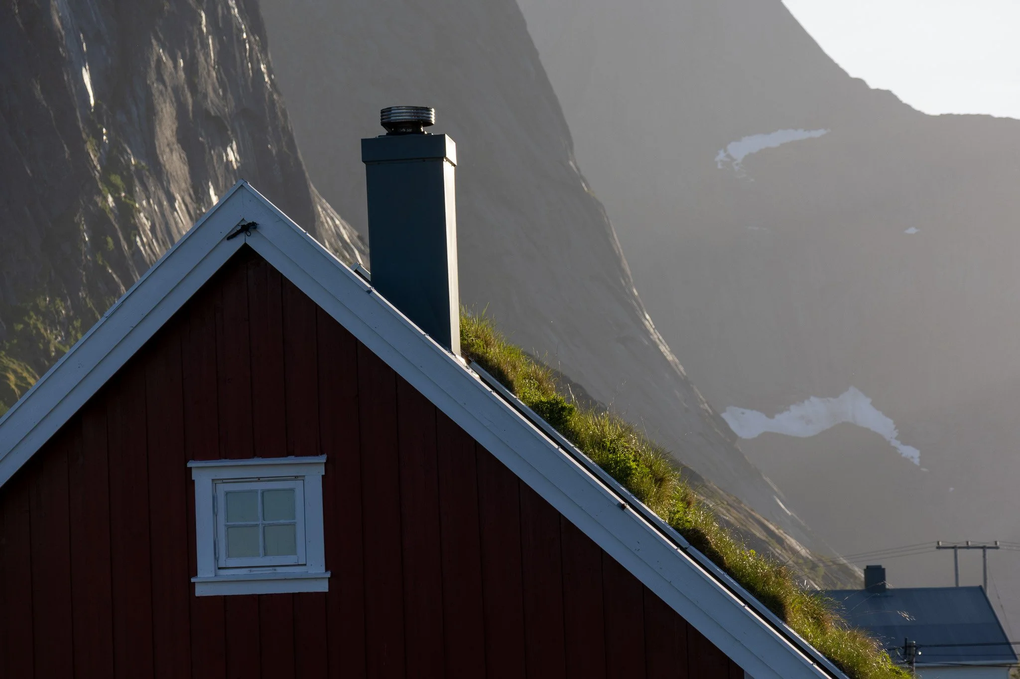 Part of a red house with a white window and a chimney on a sloped roof covered in grass. Mountain peaks are visible in the background.