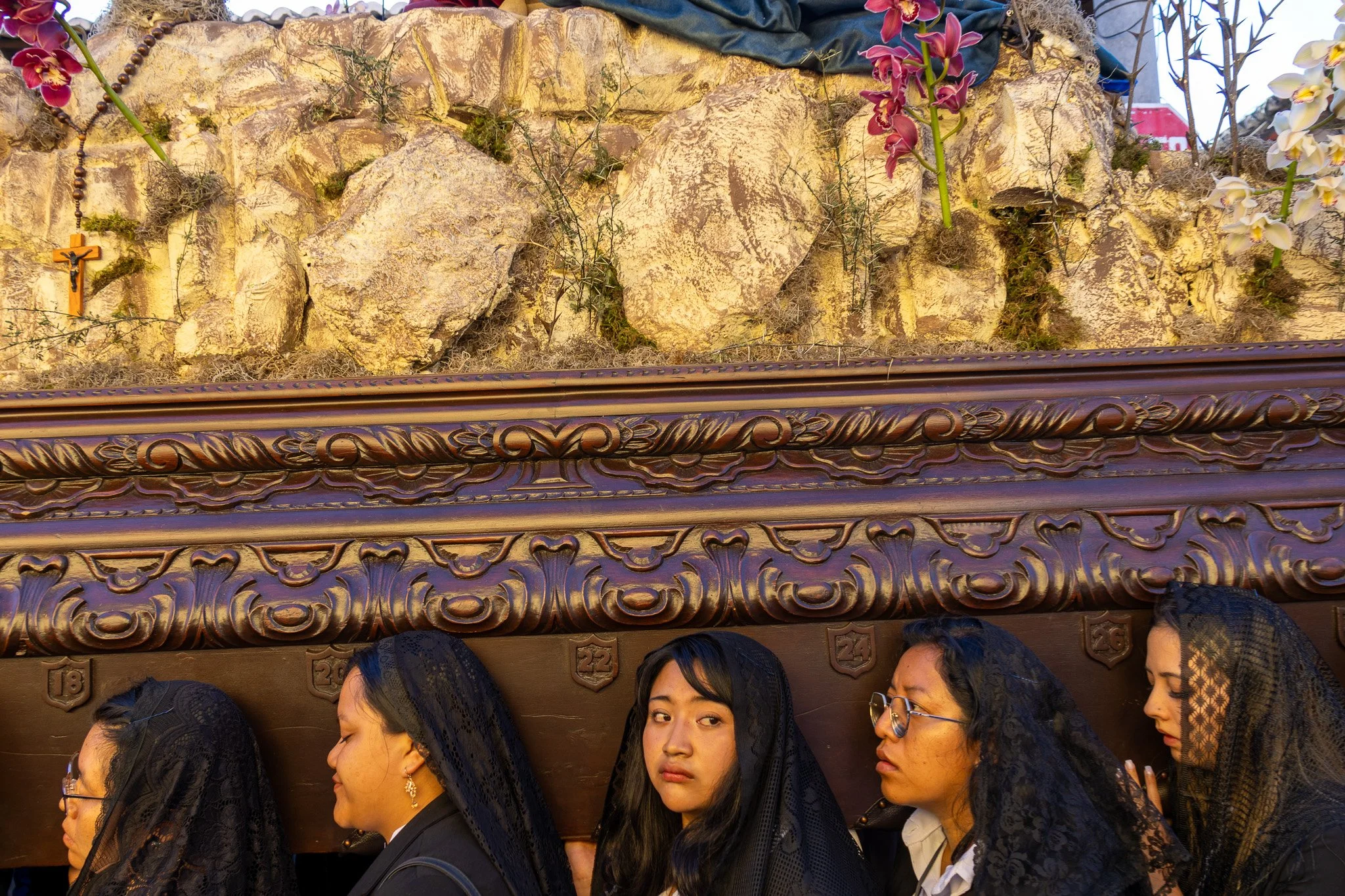 A group of women dressed in black with lace veils are walking in a religious procession, carrying a large wooden platform with carved details. The platform has decorative greens, orchids, and a rock arrangement on top.