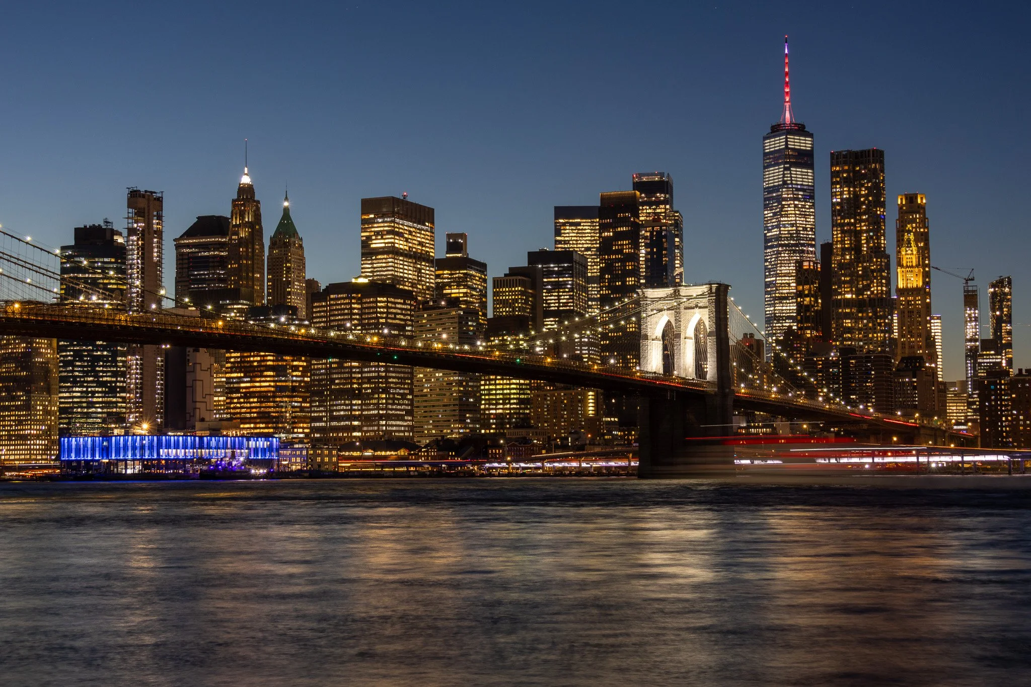 Night view of New York City skyline with illuminated skyscrapers, Brooklyn Bridge, and reflections on the water.