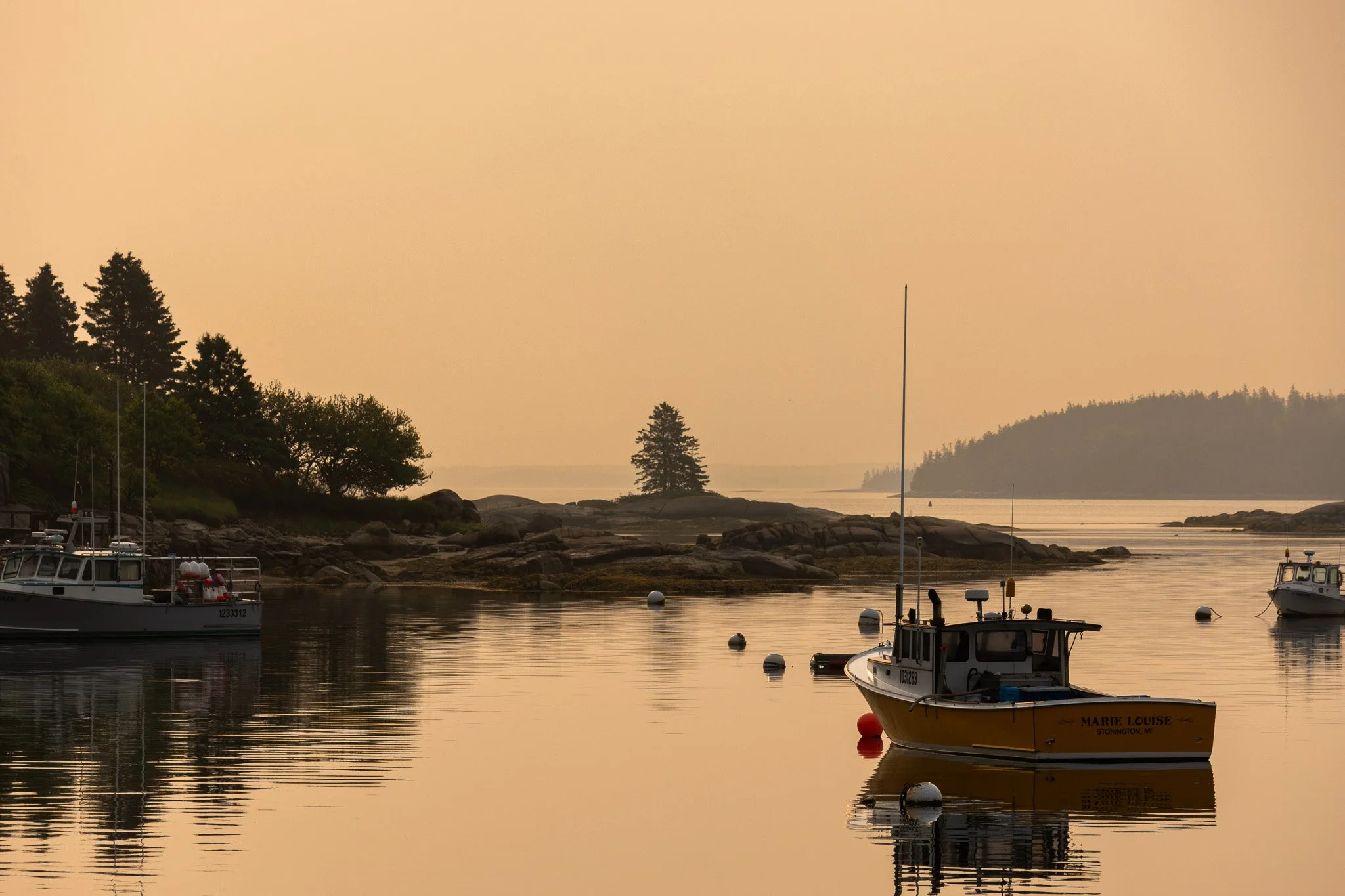 Boats anchored in calm waters near rocky shoreline at sunset, with trees and distant land in the background.