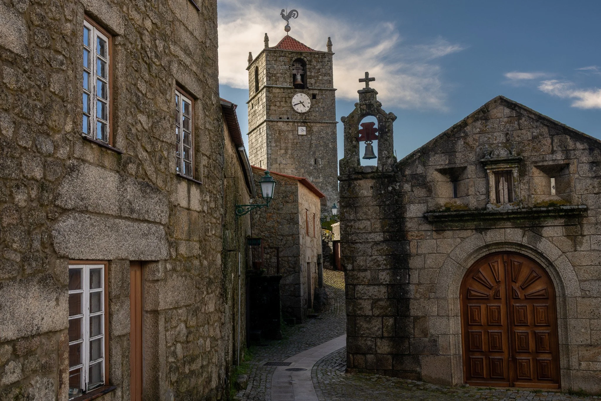 A narrow cobblestone street in a historic European village, with old stone buildings, a clock tower, and a small chapel with a wooden door and bell tower.
