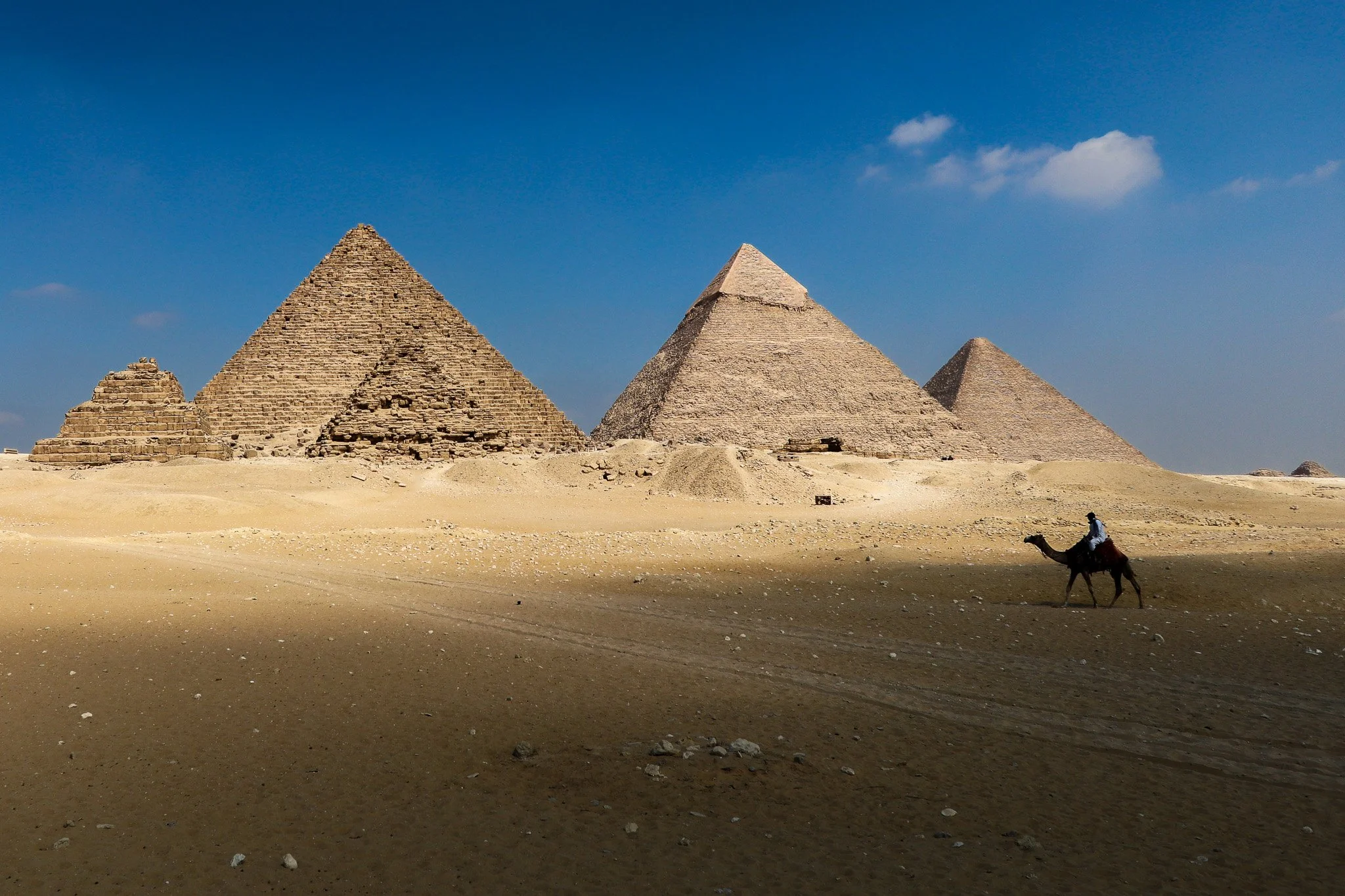 Four ancient pyramids in a desert landscape with a person riding a camel in the foreground and a blue sky above.