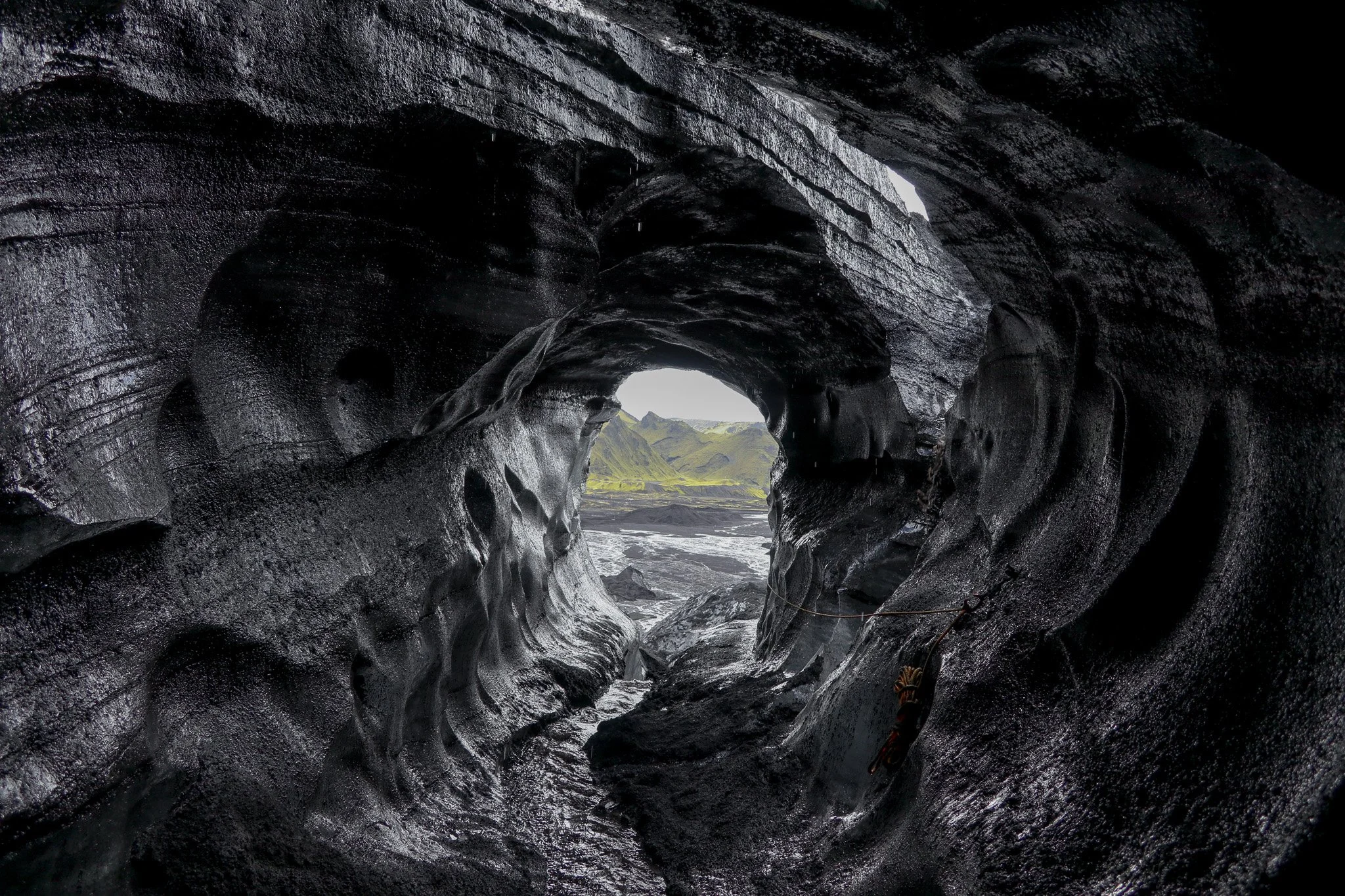 View through a dark volcanic rock tunnel towards lush green mountains and a river in the distance.