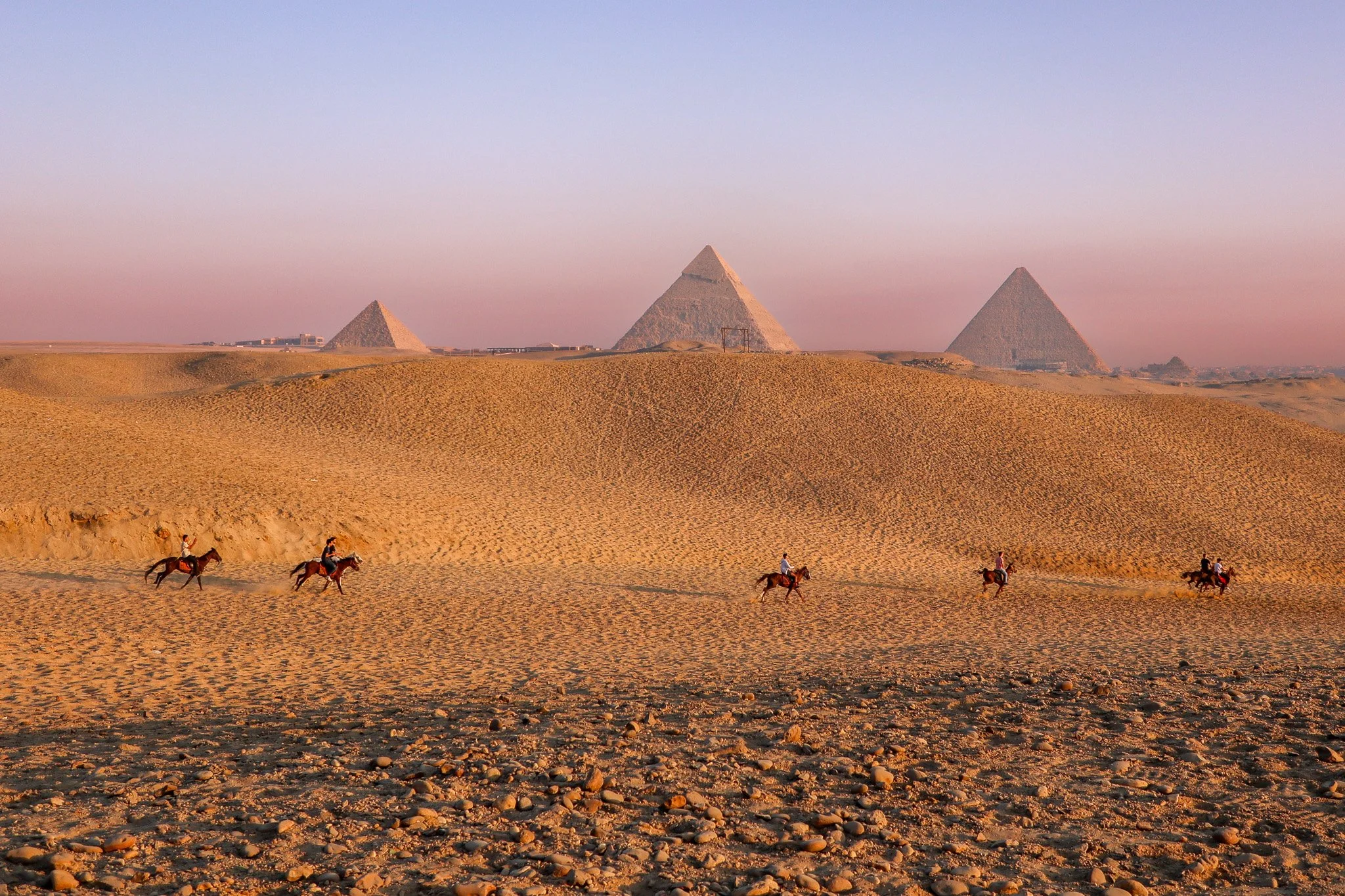 Camel riders traveling across the desert with pyramids in the background during sunset.
