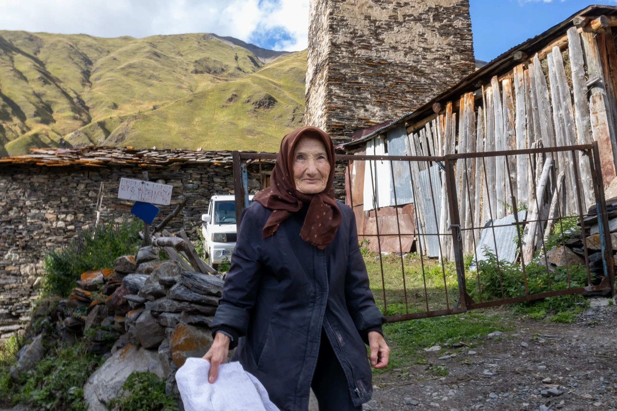 An elderly woman in a dark jacket and headscarf standing outdoors in front of rustic buildings with mountains in the background. She holds a white cloth in her hand.