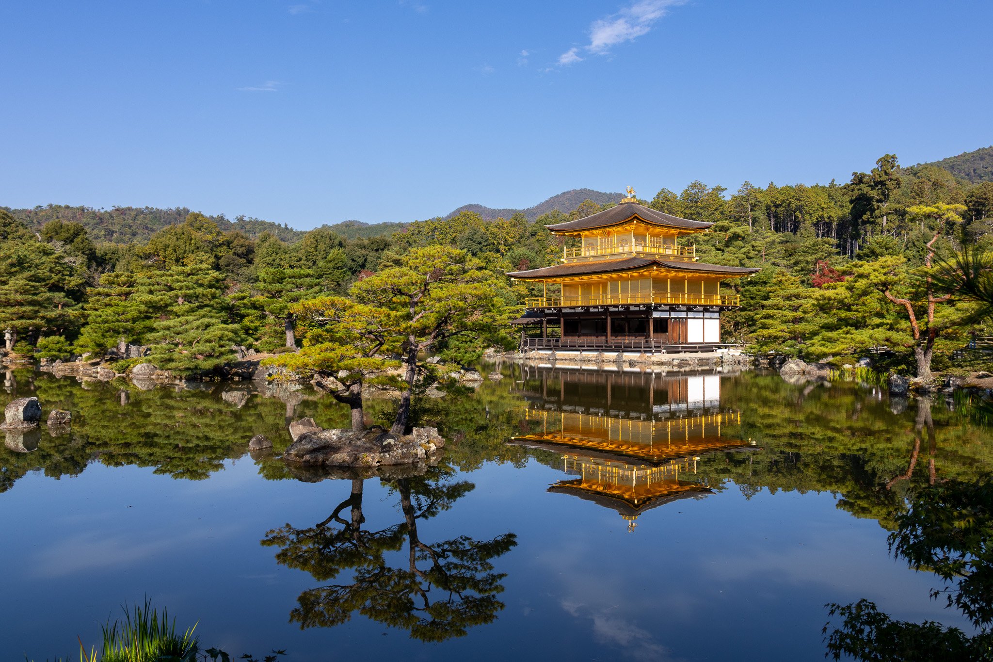 Traditional Japanese temple with gold accents reflected in a tranquil pond surrounded by lush greenery and mountains in the background.