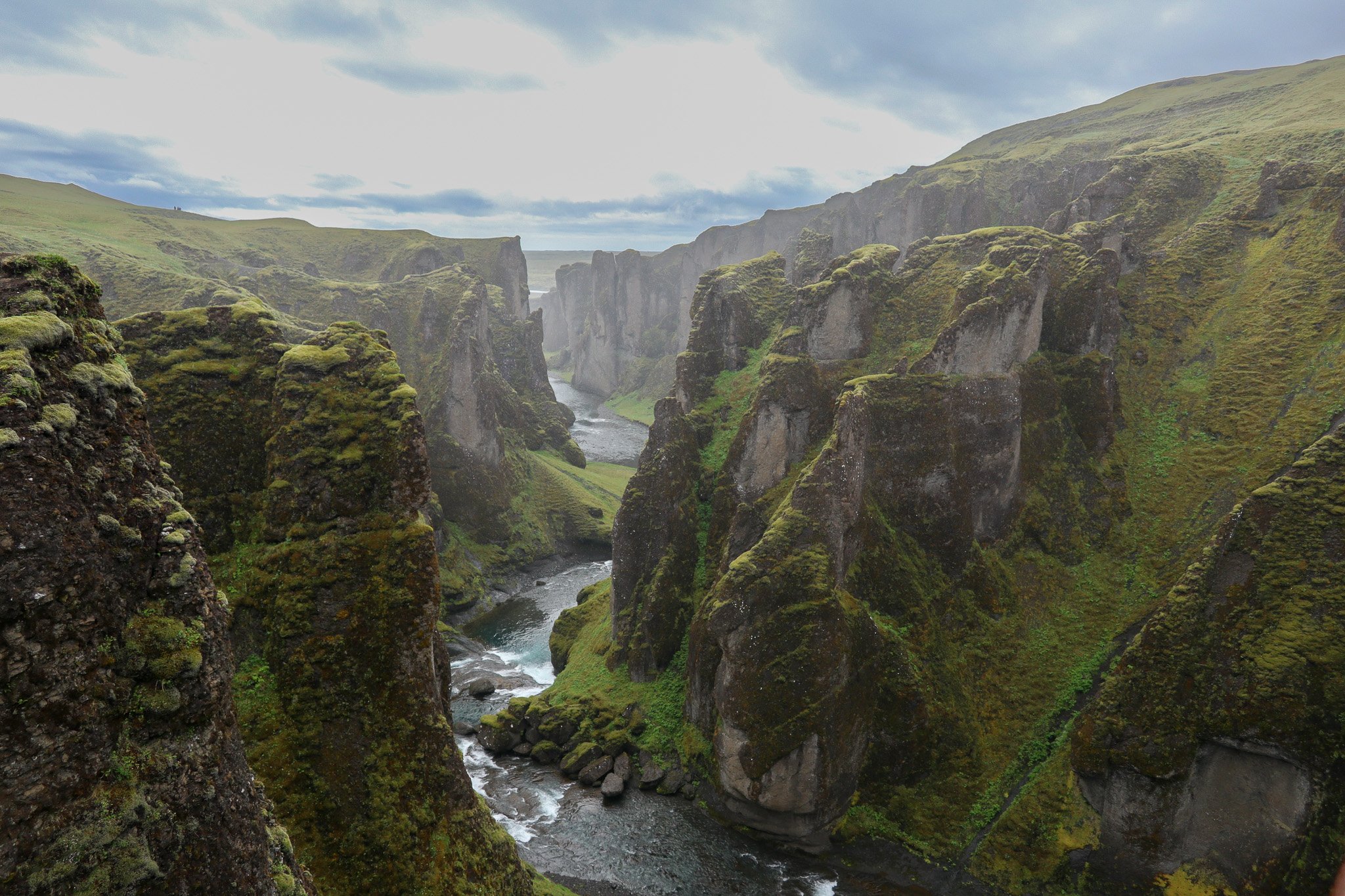 Moss-covered cliffs and a river flowing through a canyon in a lush, green landscape under a cloudy sky.