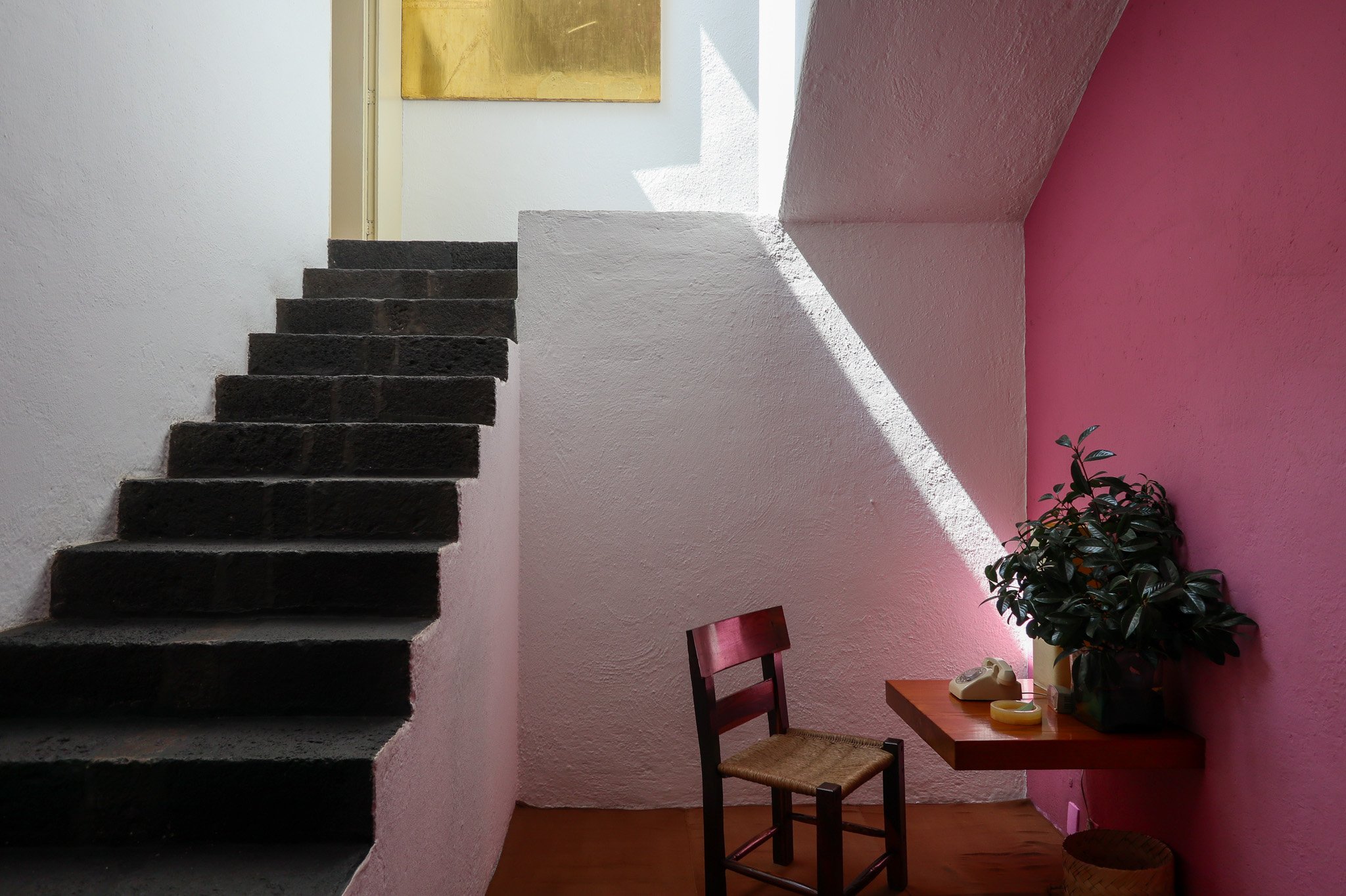 Interior of a pink and white room with a set of dark stairs, a wooden chair, a small wall-mounted wooden table with a potted plant, a white rotary phone, and a yellow ceramic object with sunlight casting shadows.