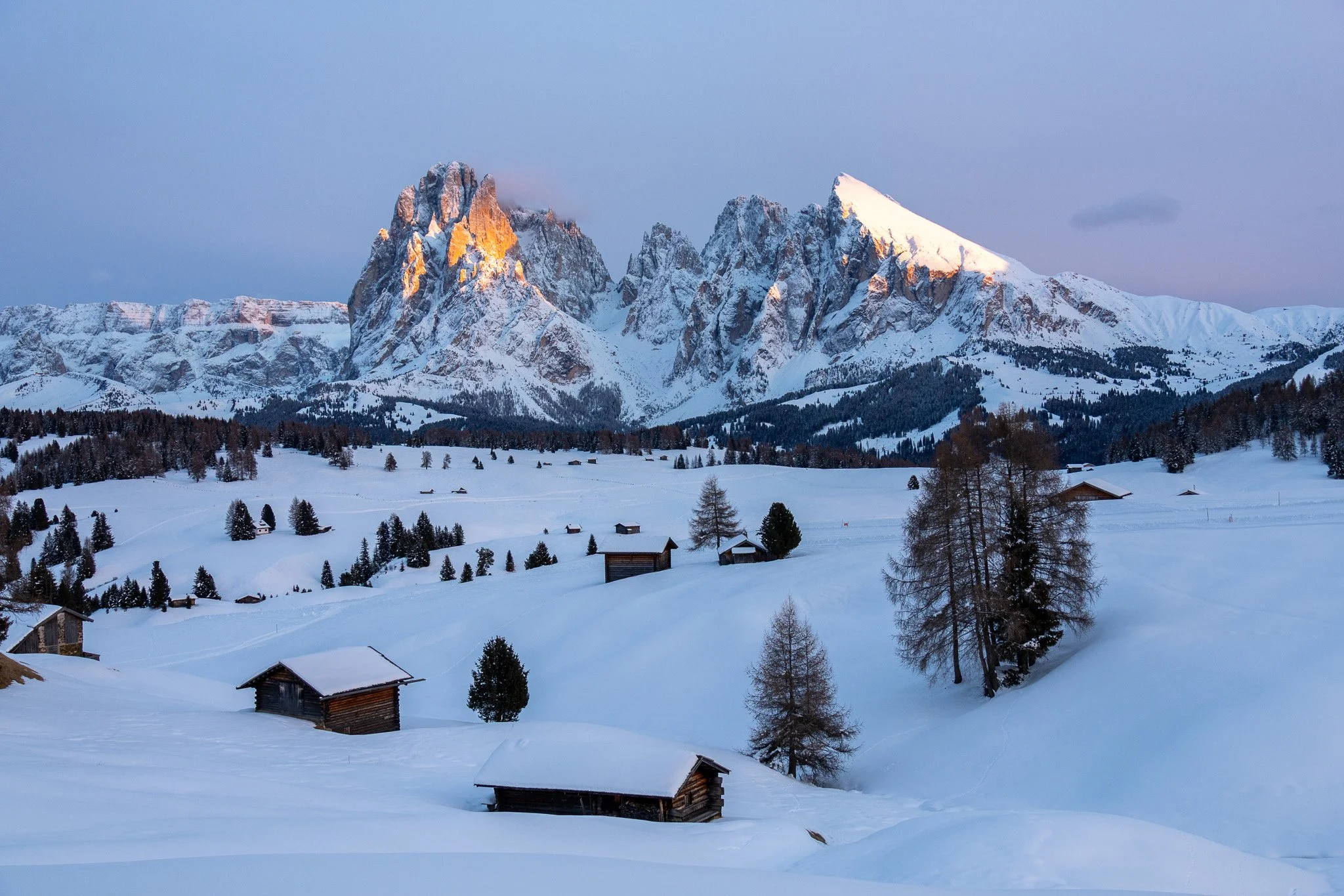 Snow-covered landscape with small wooden cabins and trees leading up to a mountain range, likely during dawn or dusk.
