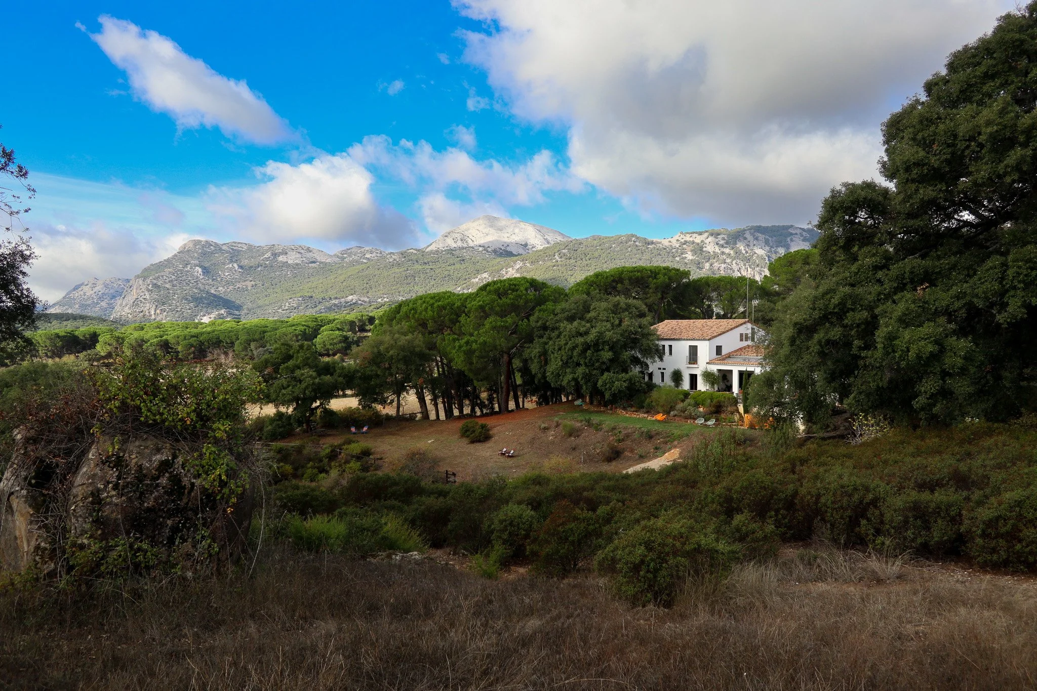 A white house with a tiled roof surrounded by green trees and hills with mountains in the background under a partly cloudy sky.