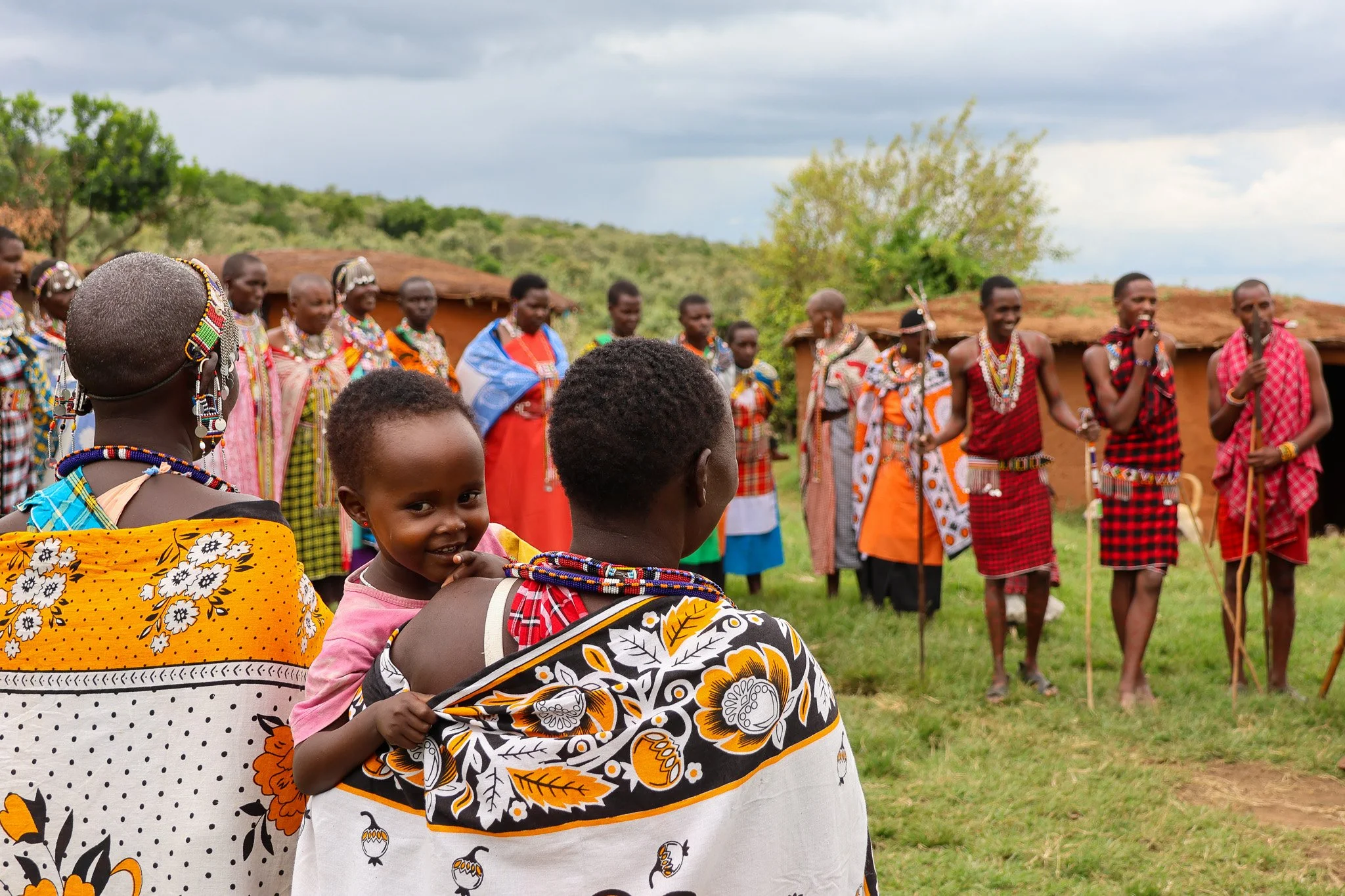 A group of Maasai people wearing traditional clothing and jewelry, gathered outdoors in a rural area with grass and small huts, participating in a cultural event or dance.