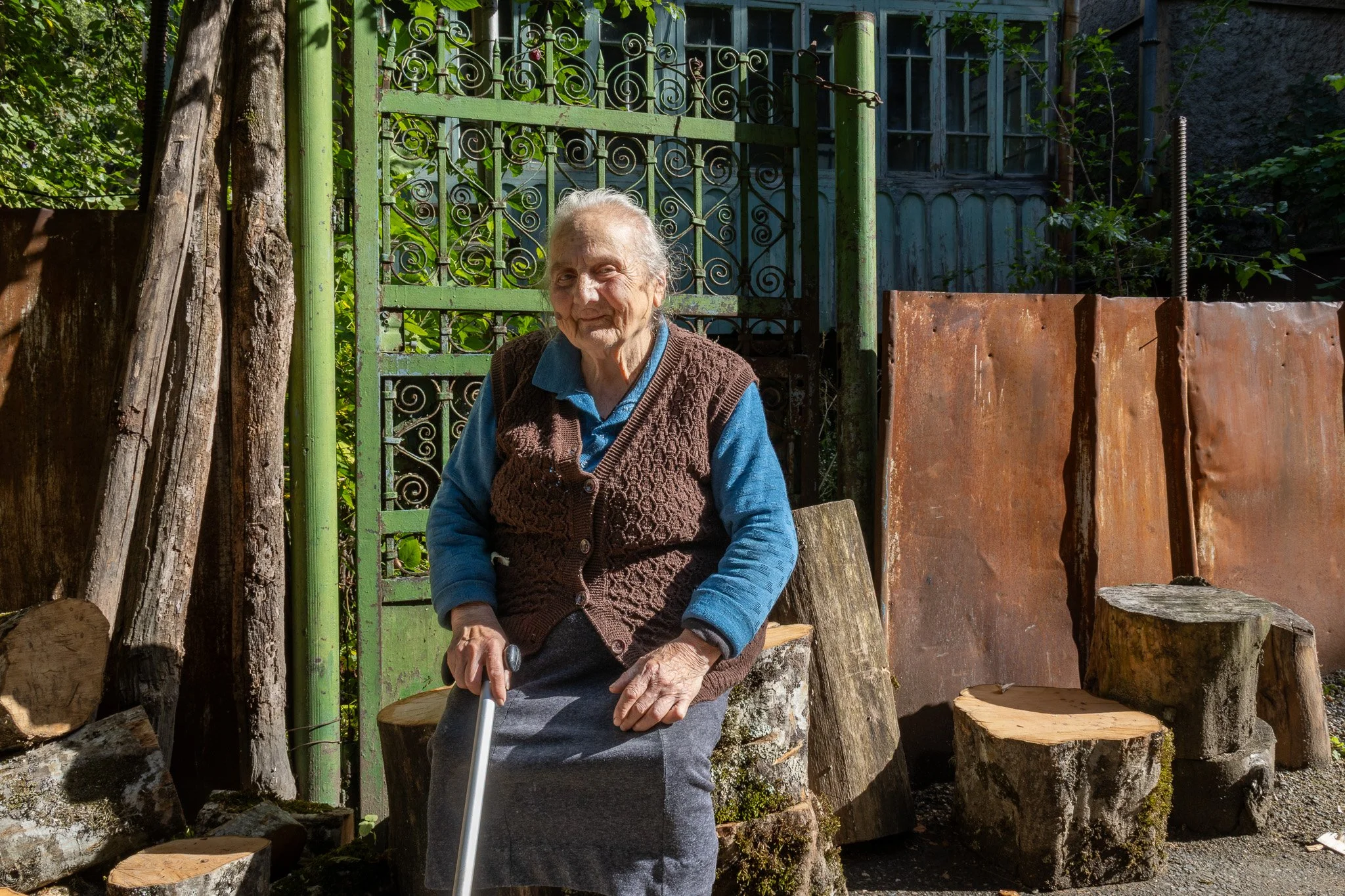 An elderly woman sitting outdoors on a piece of wood, holding a cane, with a rustic, green metal gate and natural wood logs behind her.