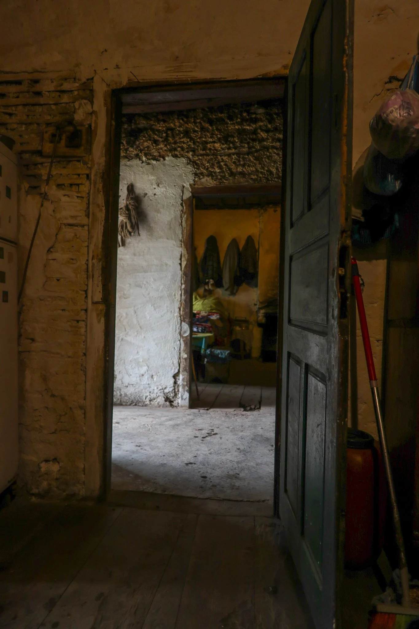 View through a rustic, open doorway into a dimly lit interior with exposed brick and plaster walls, showing coats hanging on the wall and various household items.