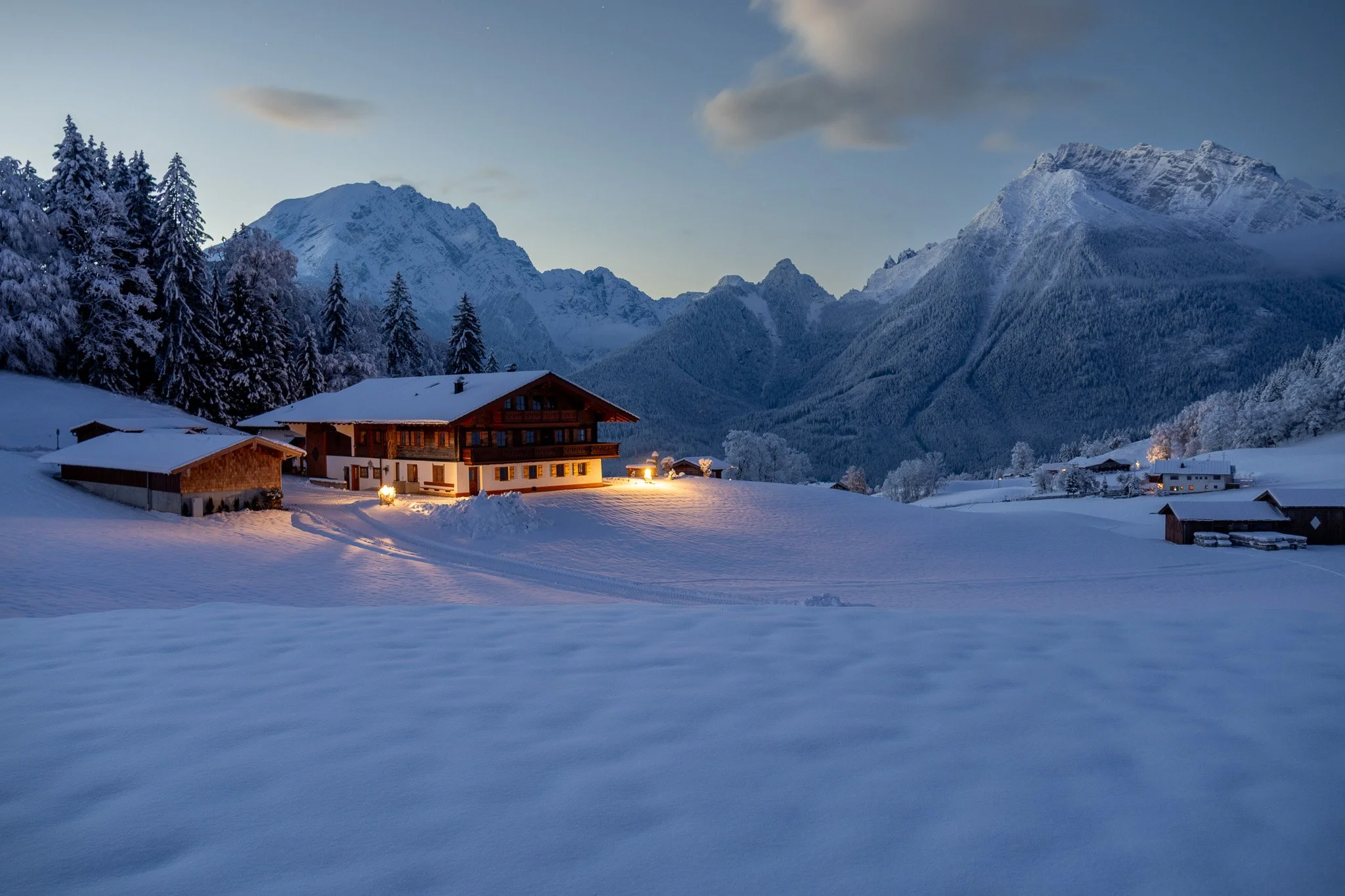 Snow-covered landscape with a chalet-style house and other buildings, surrounded by pine trees and mountains in the background, at dusk.