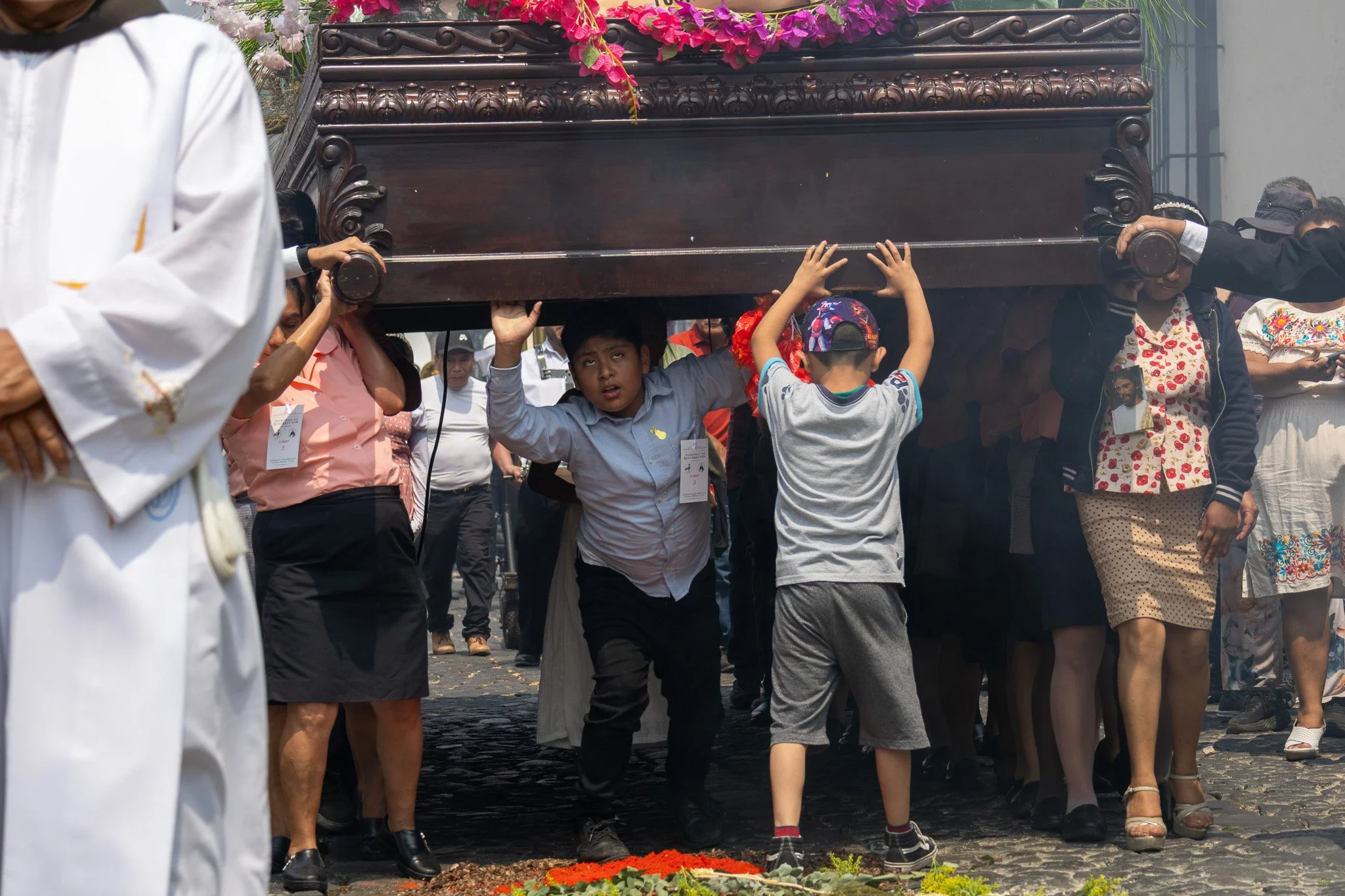 People participating in a procession, carrying a wooden casket at a religious or cultural event. The scene is outdoors on a cobblestone street with floral decorations.