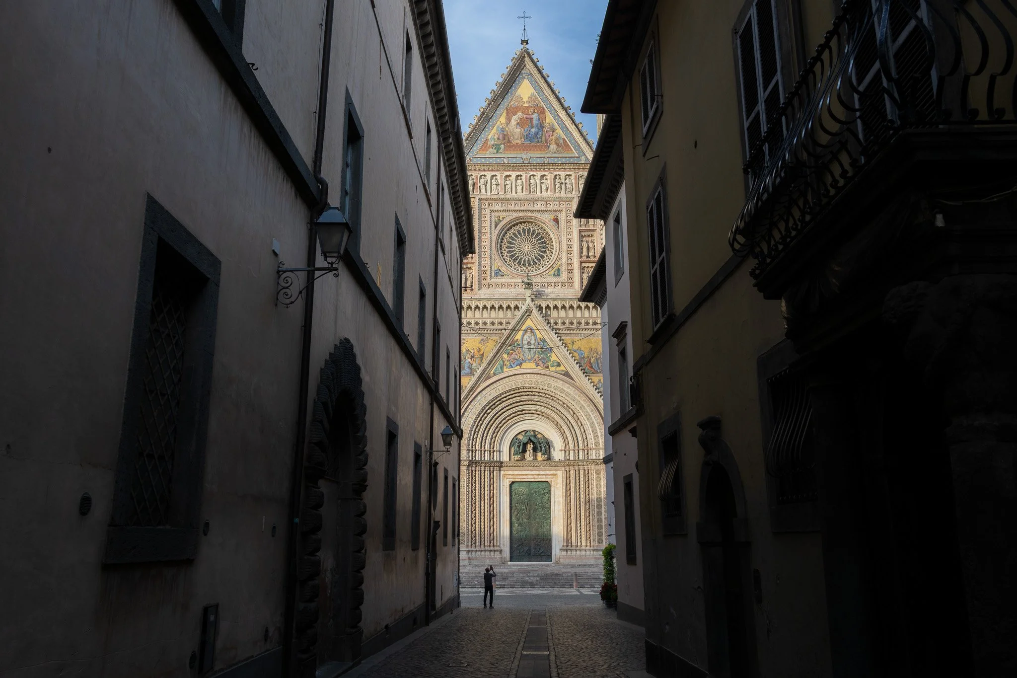 View of Florence Cathedral's bell tower, seen from a narrow alleyway with buildings on both sides, and a person taking a photo in the distance.