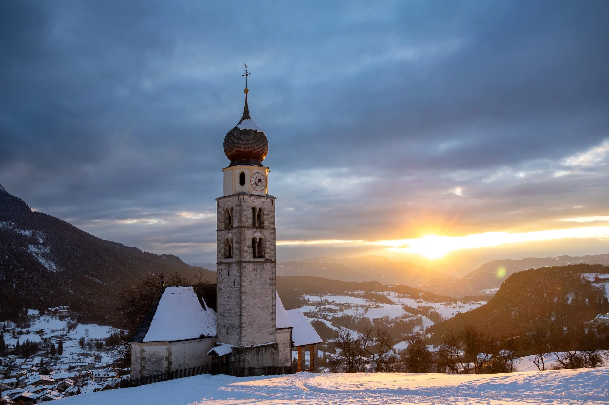 A winter landscape featuring a church with a tall bell tower, snow-covered roofs, and surrounding hills at sunset with cloudy sky.