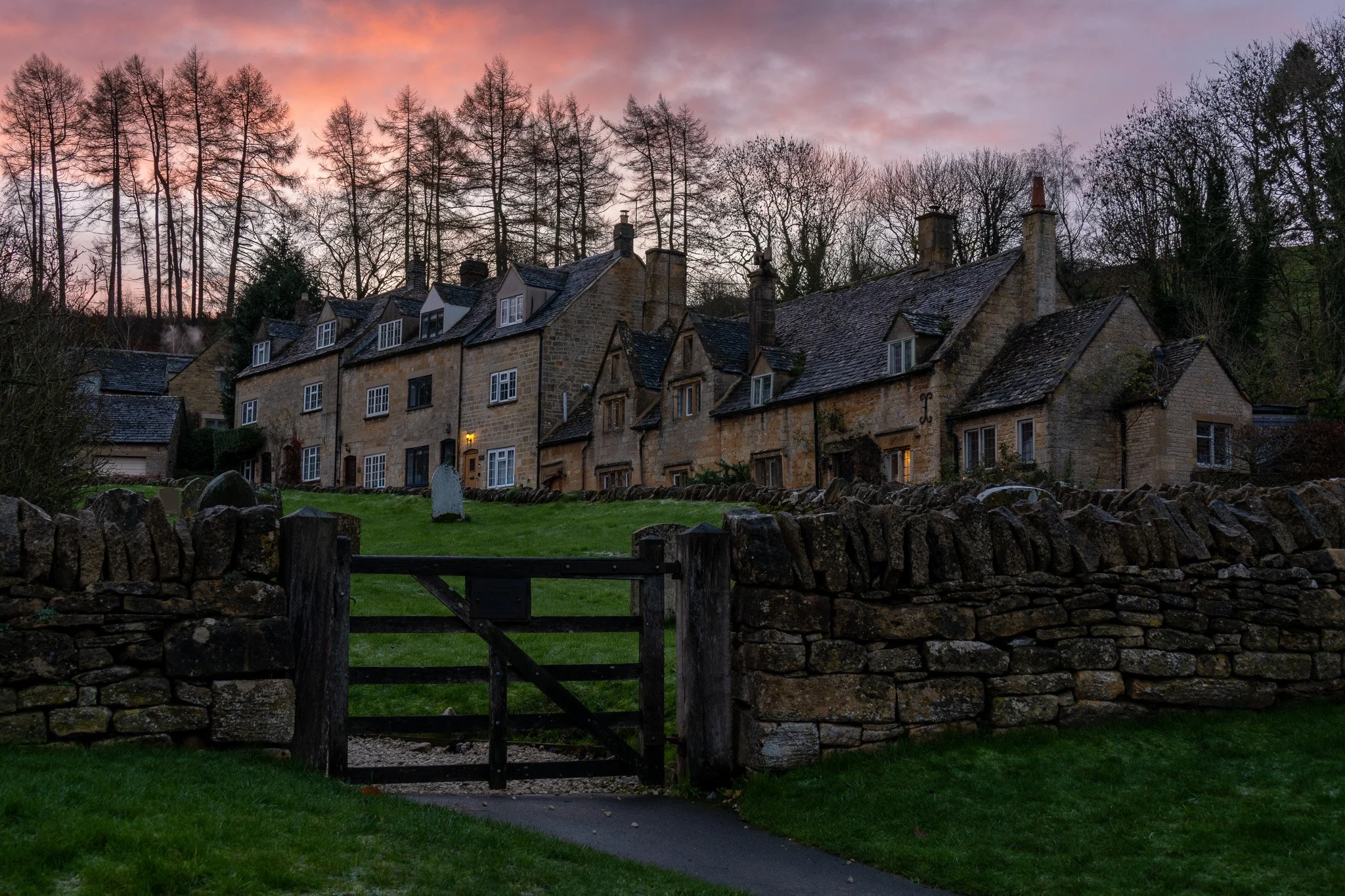 A row of old stone houses with slate roofs and chimneys, surrounded by a stone wall and a grassy yard, during sunset with pink and purple sky.