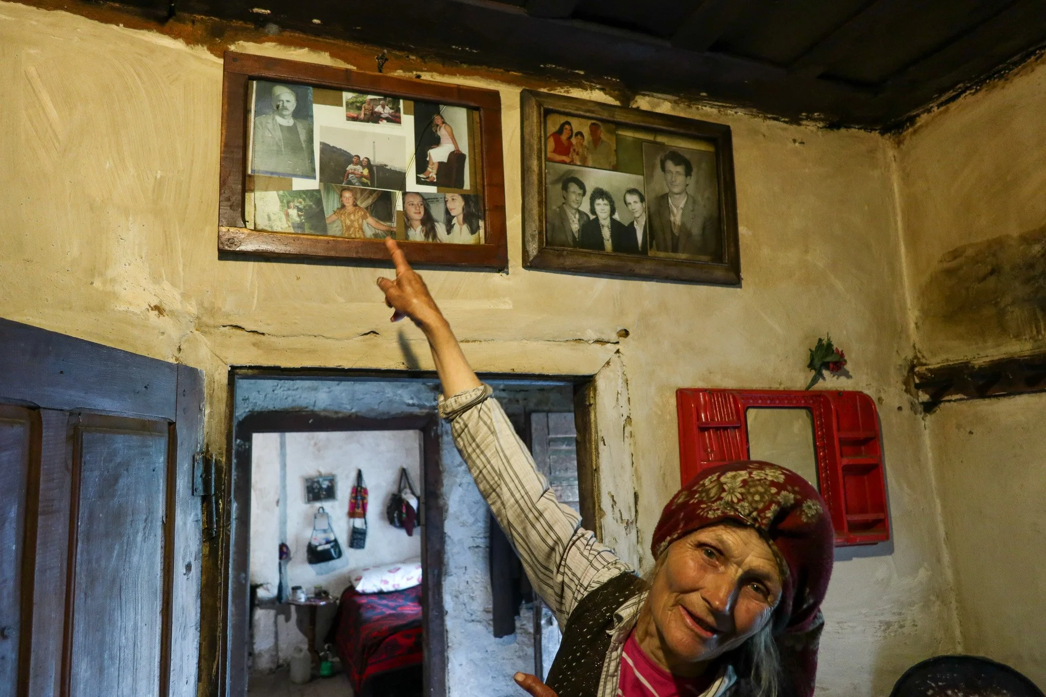 An elderly woman wearing a headscarf points to a collage of old photographs in wooden frames on a beige wall inside a rustic home, with a glimpse of a bedroom in the background.