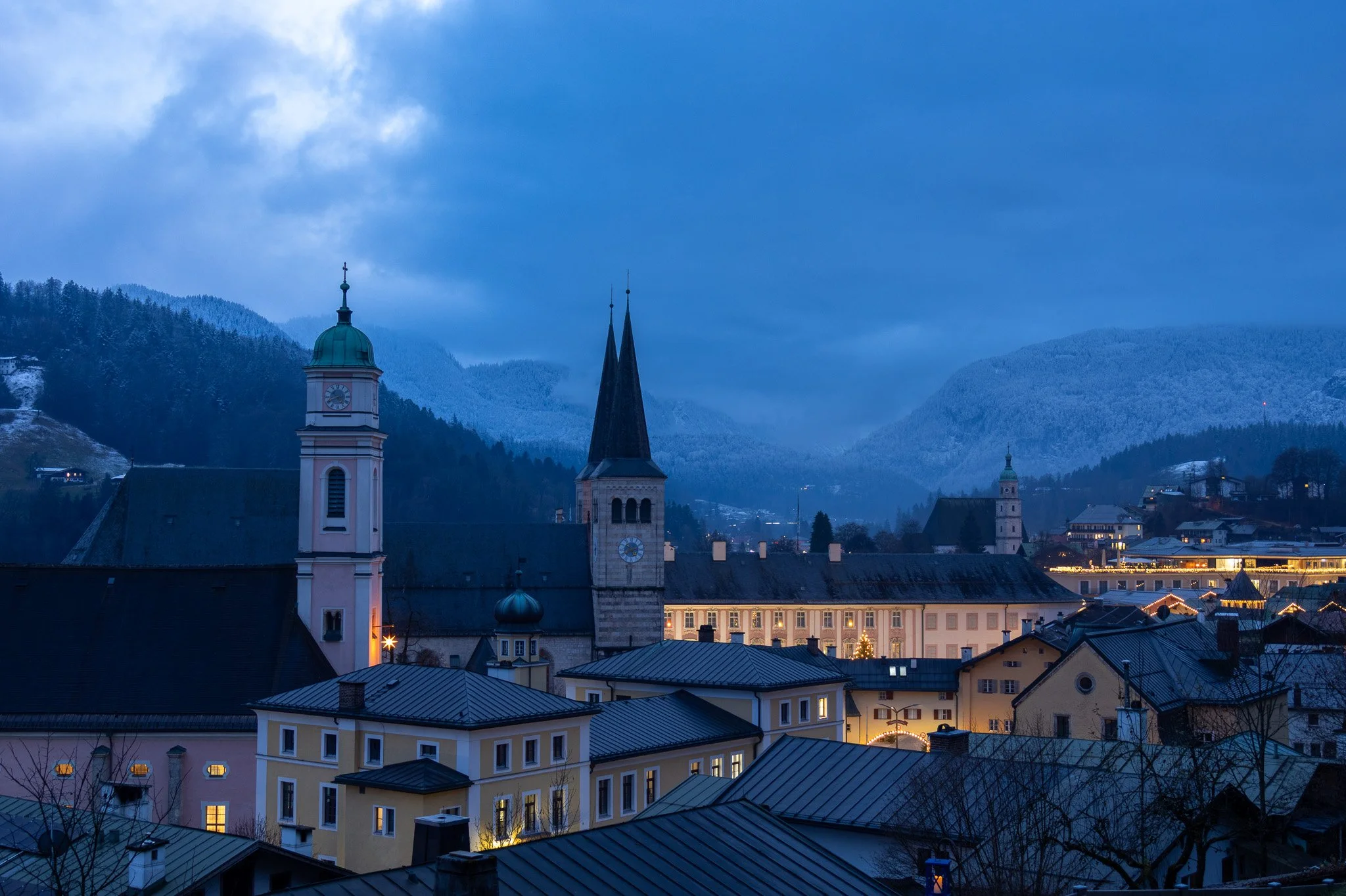 View of a European town with church steeples and buildings illuminated at dusk, surrounded by snow-covered mountains.