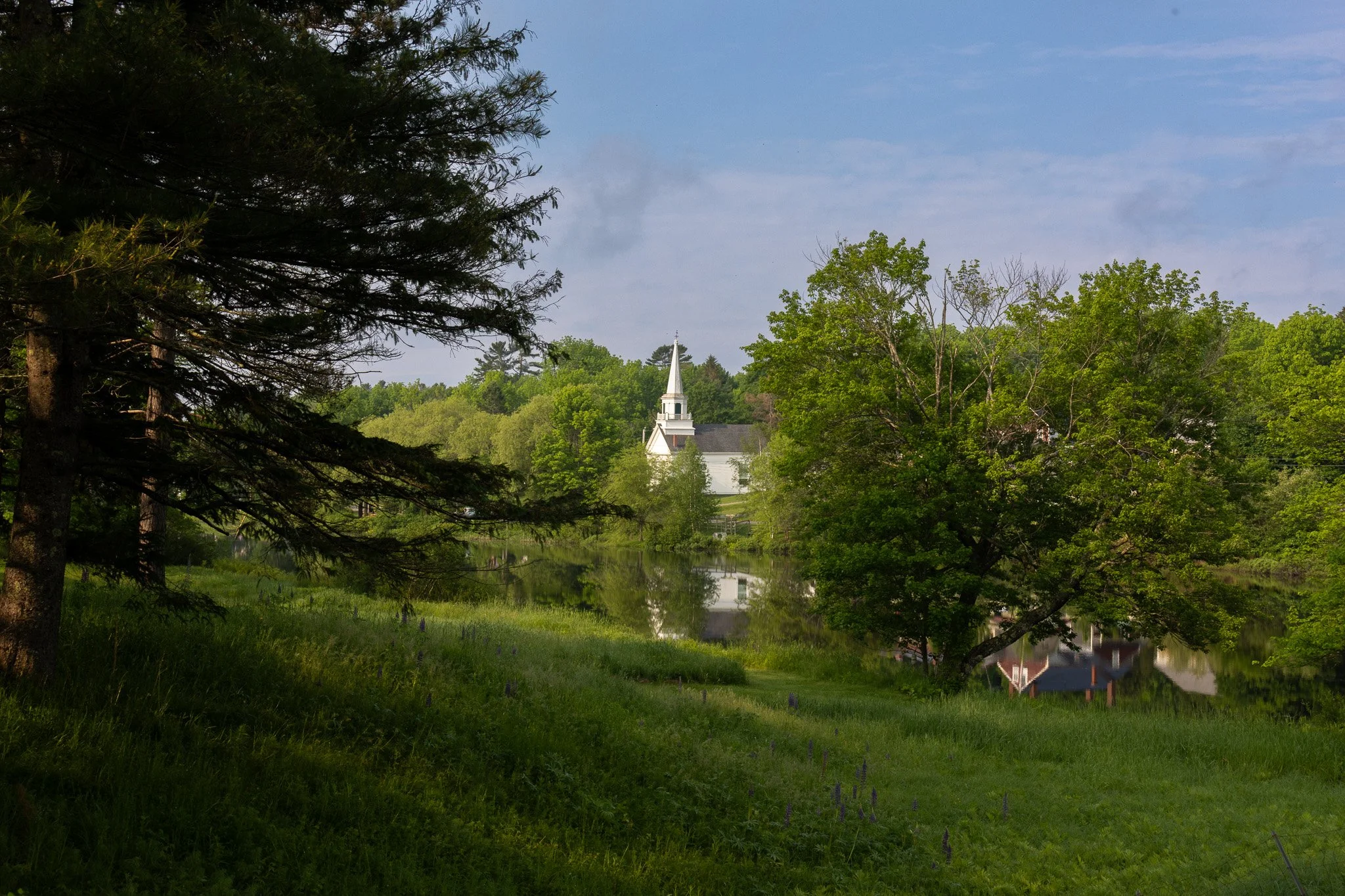 A serene landscape with lush green trees and grass, a body of water reflecting a white church with a tall steeple, and a partly cloudy sky.
