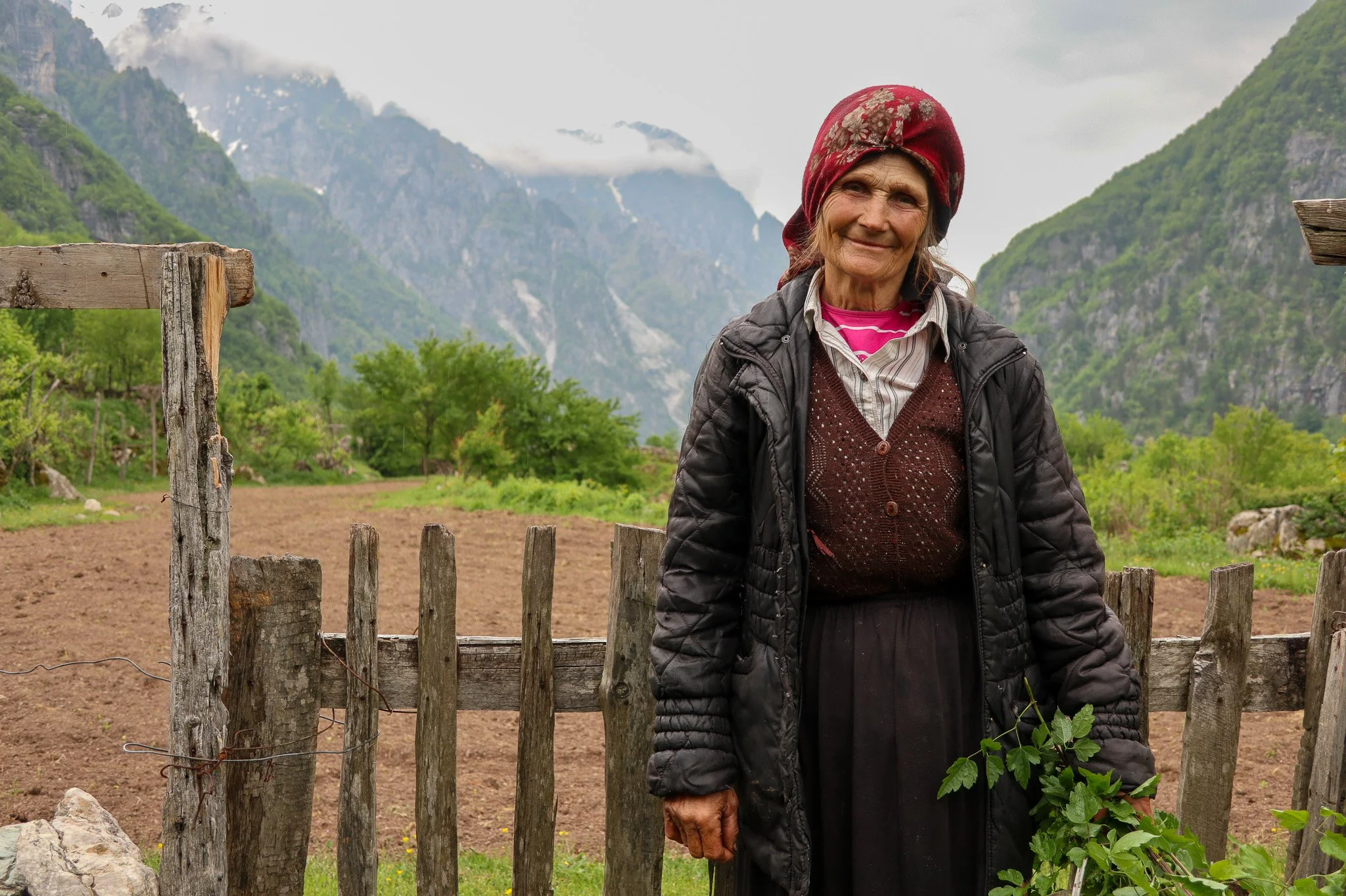 An elderly woman with gray hair, wearing a red headscarf, a dark jacket, a sweater, and a striped shirt, standing outdoors near a wooden fence in a rural area with mountains and green trees in the background.