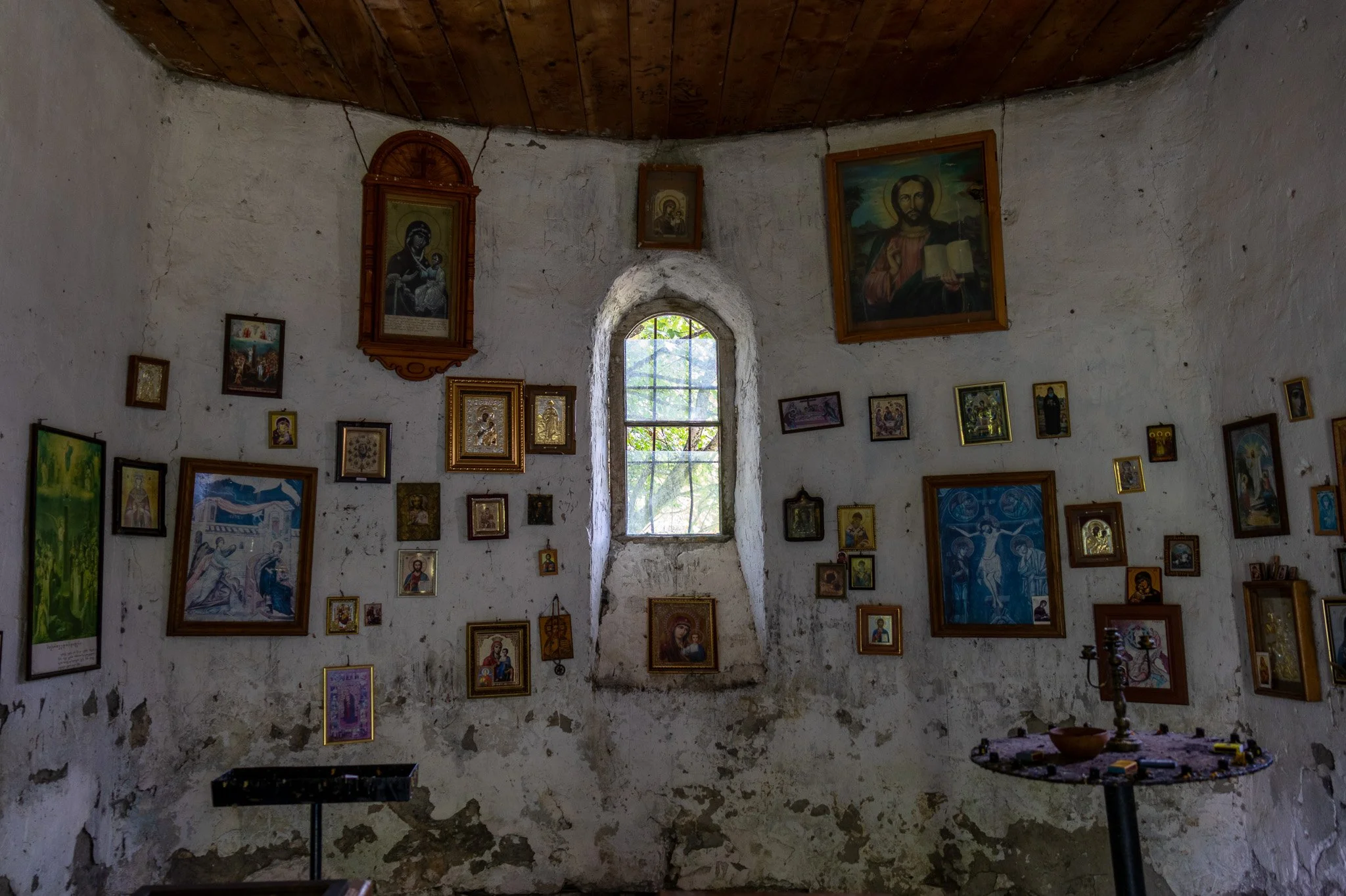 Interior of a small chapel with white plastered walls and a curved wooden ceiling, decorated with numerous framed religious icons and paintings, a small arched window in the center letting in natural light.