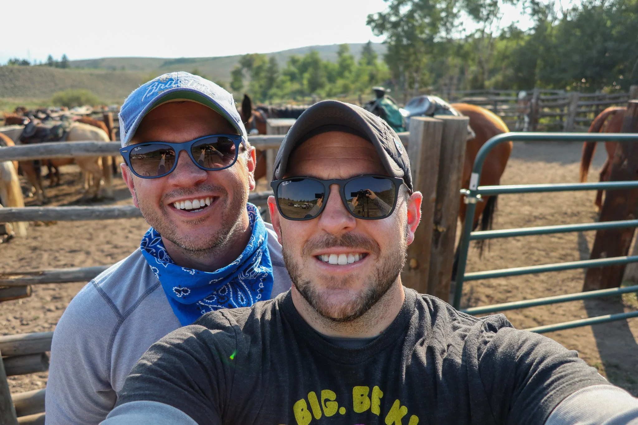 Two smiling men wearing sunglasses taking a selfie at a ranch with horses and a wooden fence in the background.