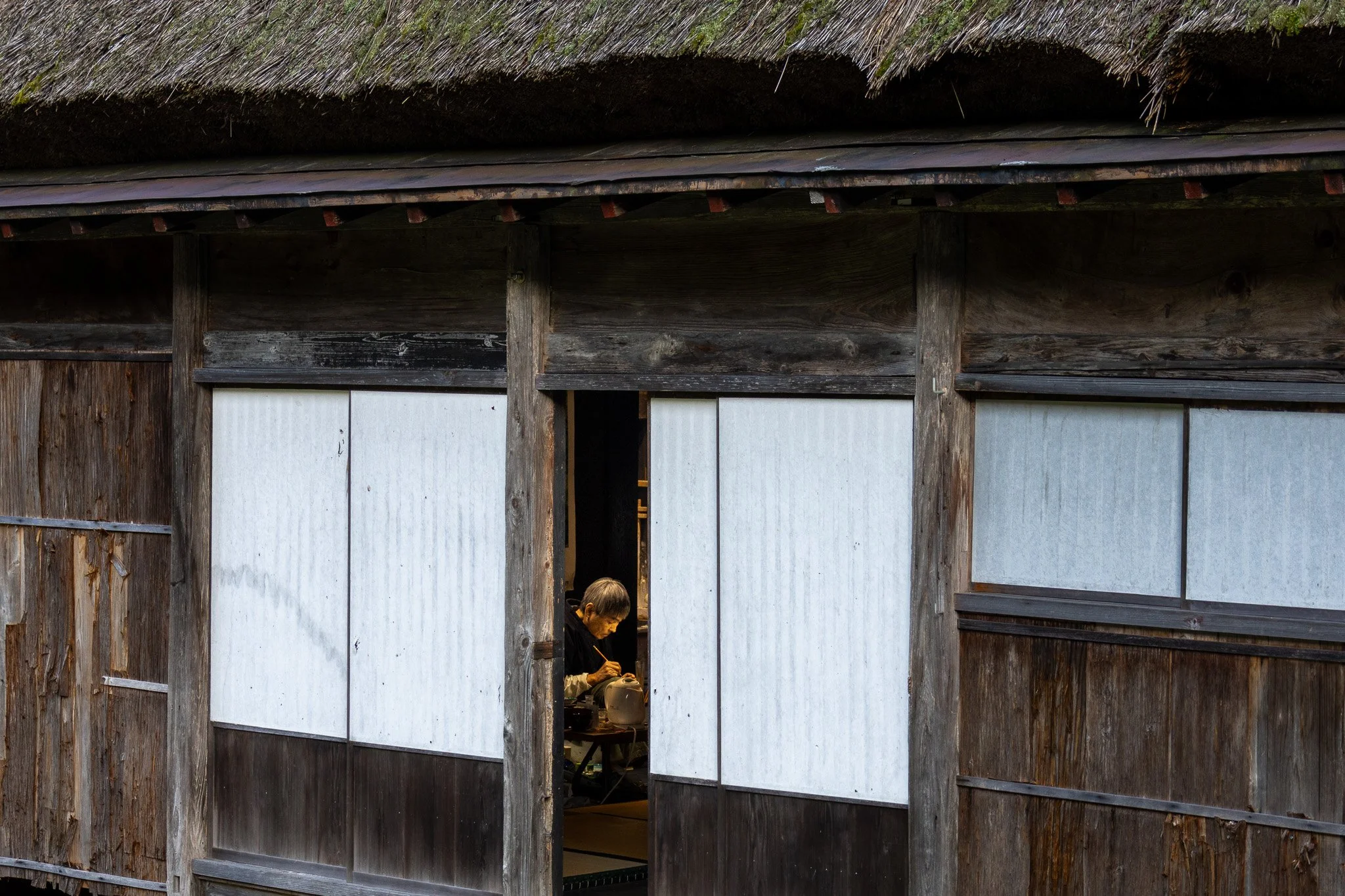 A person working inside a traditional Japanese thatched-roof house, seen through an open doorway with wooden framing and white paper panels.