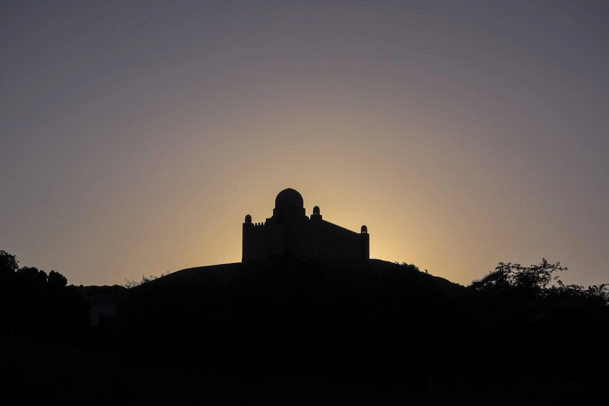 Silhouette of a historic building with domed roof on a hill, backlit by the setting or rising sun, with a clear sky in the background.