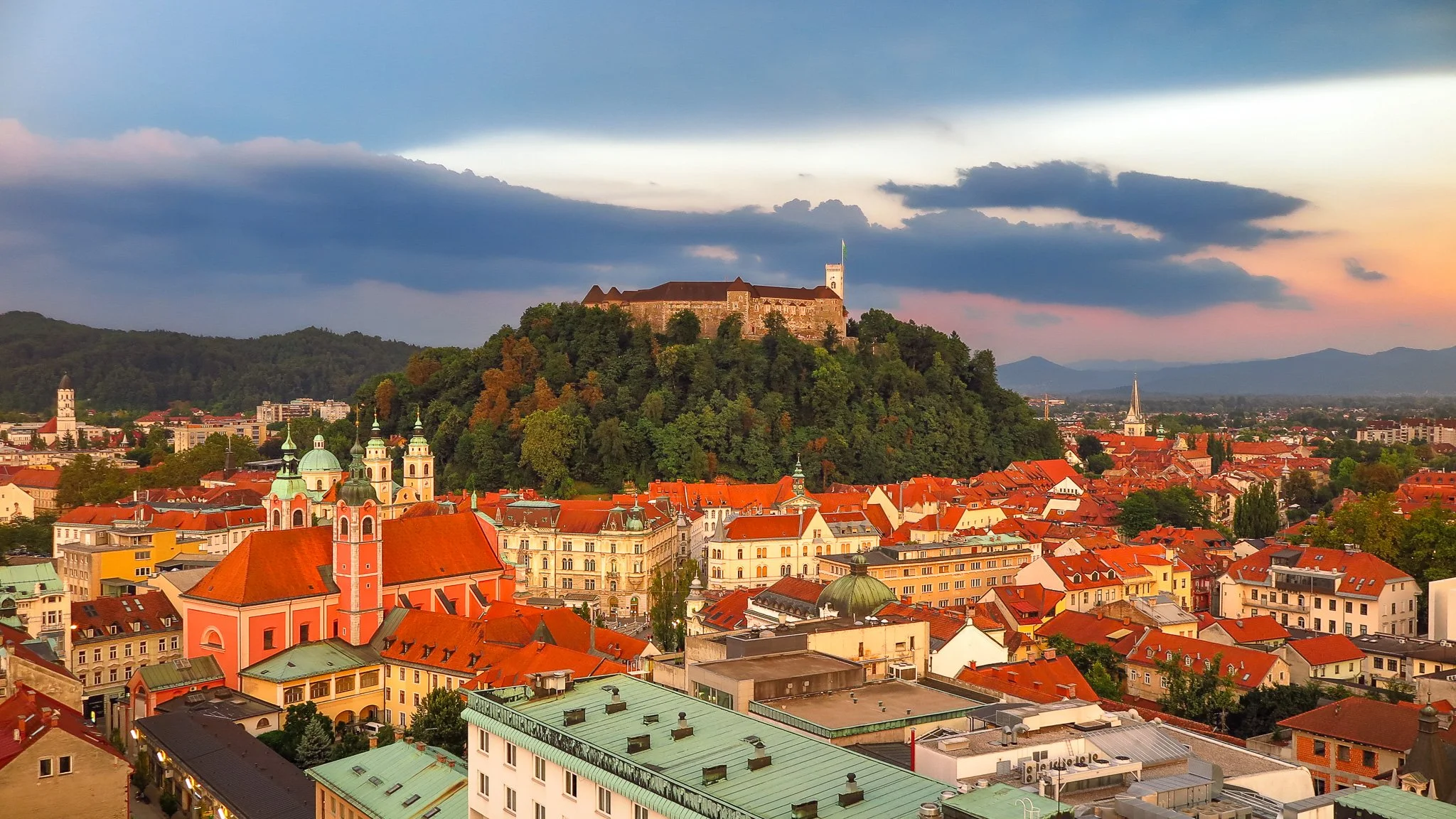 Cityscape of a European city with historic buildings, church steeples, and a castle on a hill at sunset.
