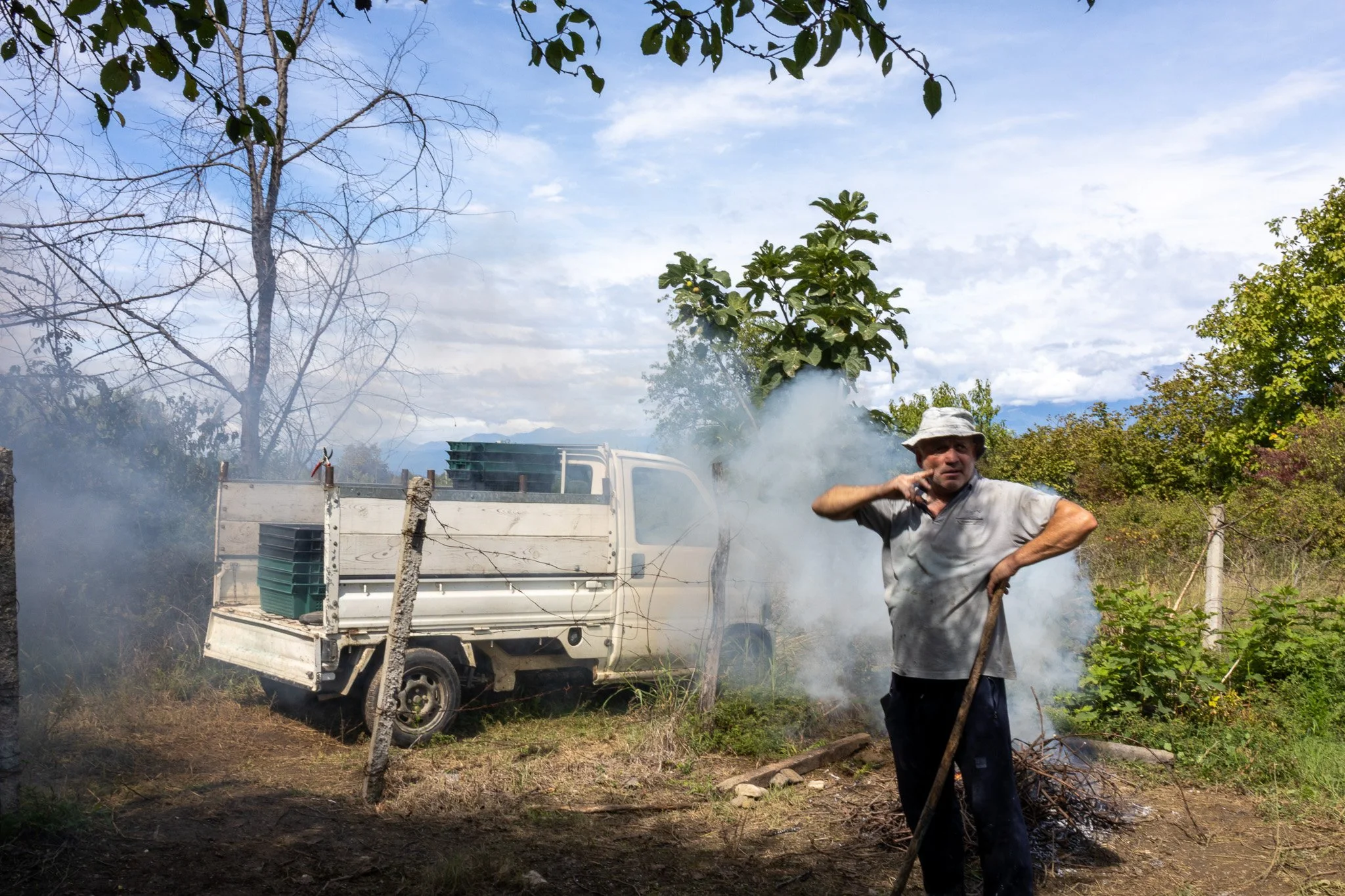 A man wearing a light-colored t-shirt, dark pants, and a wide-brimmed hat stands outdoors, holding a stick, with smoke rising from a small fire on the ground near him. Behind him is a white pickup truck loaded with plastic crates, with a rural settin