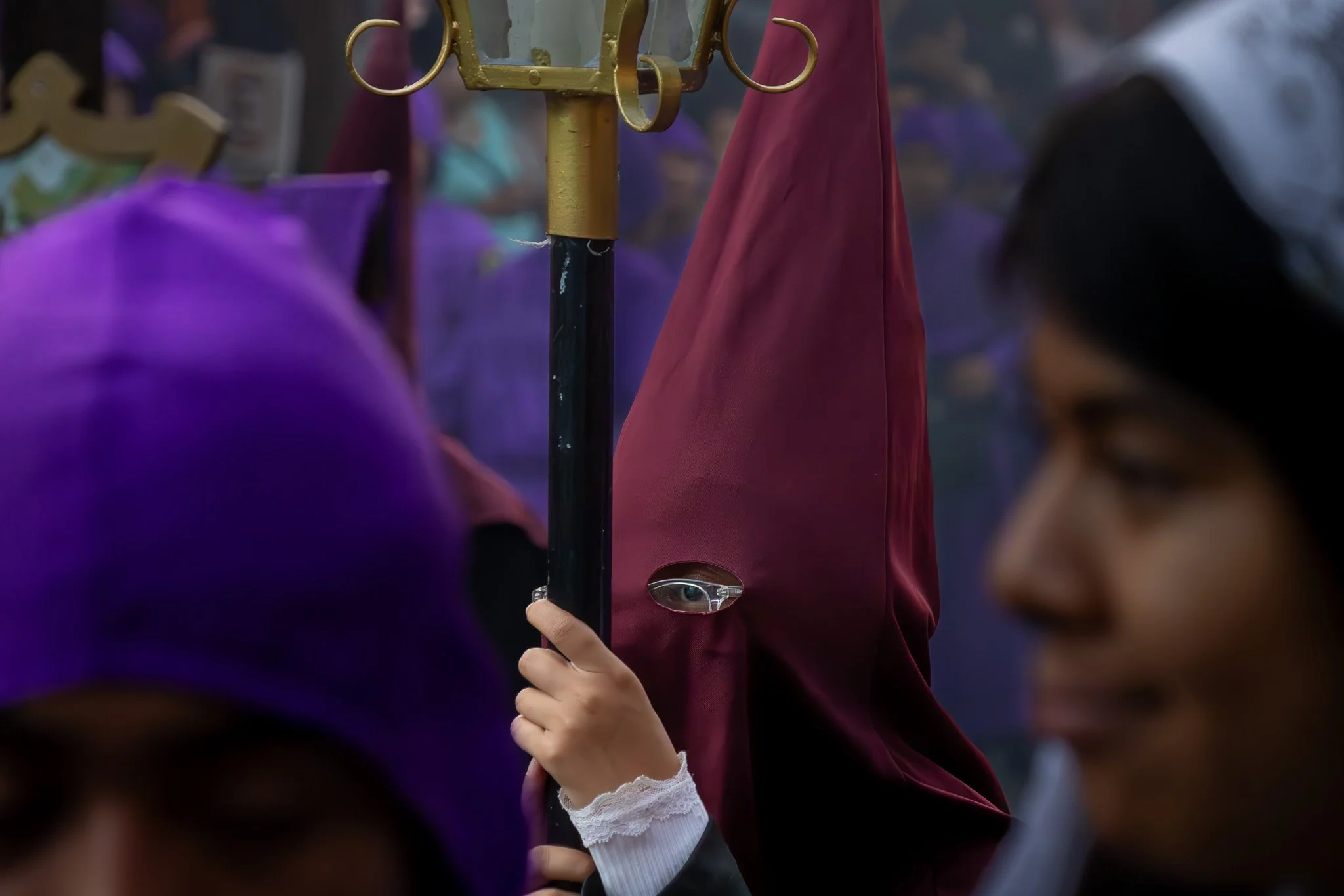 A person dressed in a hooded maroon robe with a small opening over the eye, holding a staff during a procession or festival, surrounded by other individuals, some wearing purple head coverings.