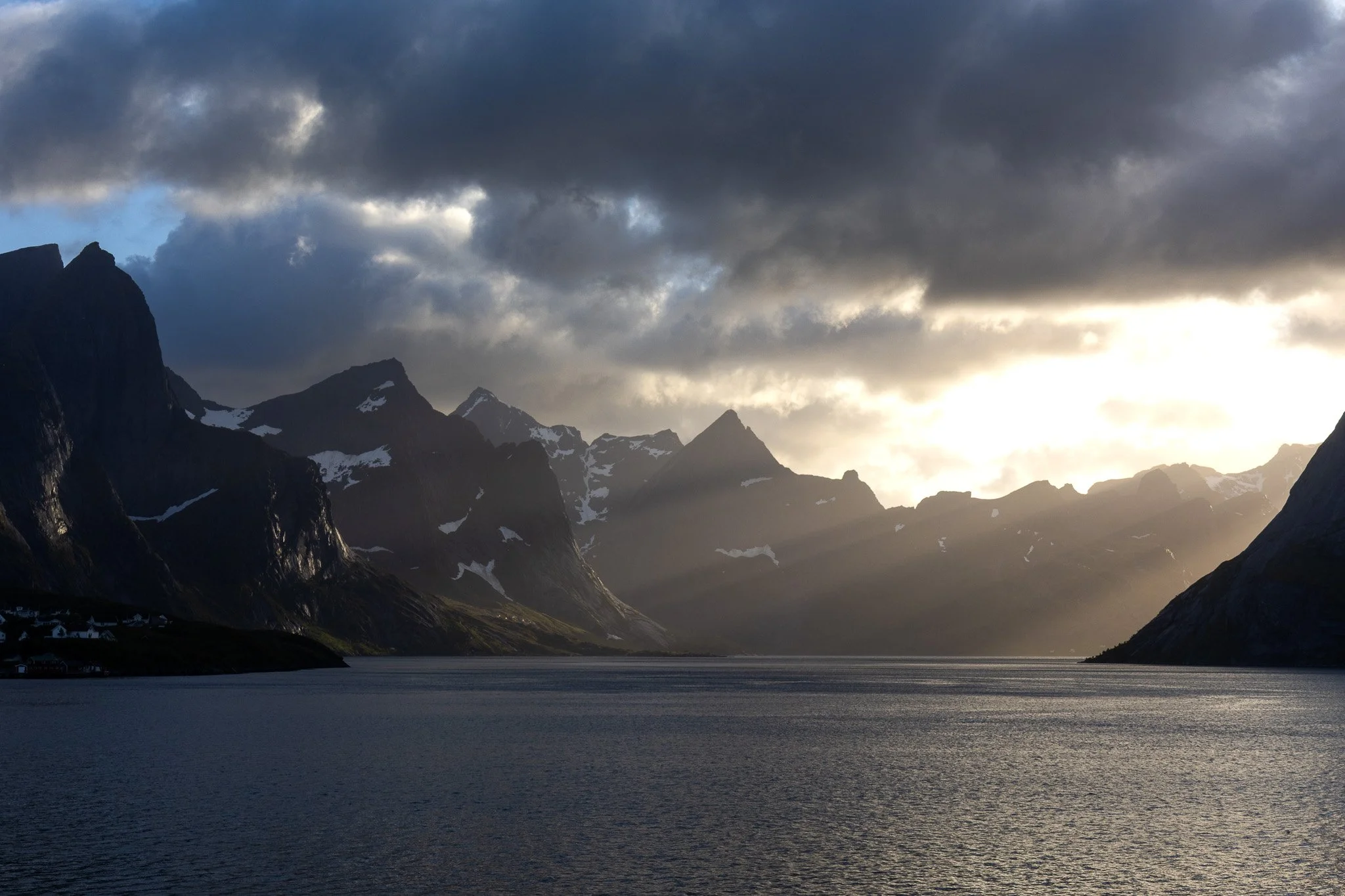 Sunset over a fjord with rugged, snow-capped mountains and dark clouds in the sky, reflecting on the water.