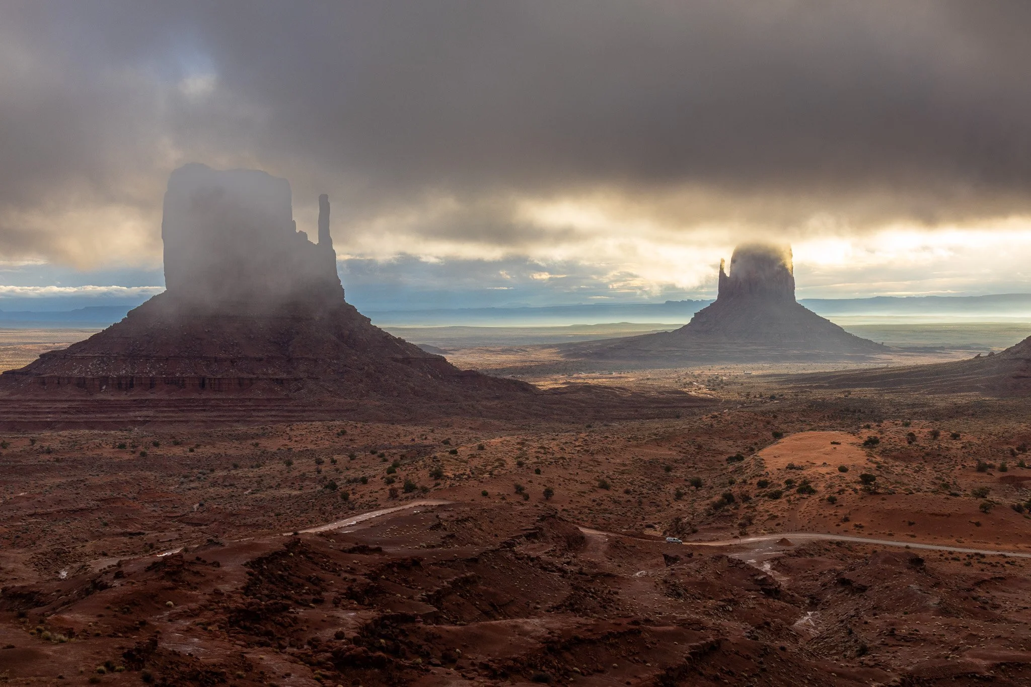 Two large rock formations in a desert landscape with dark clouds overhead and sunlight breaking through near the horizon.