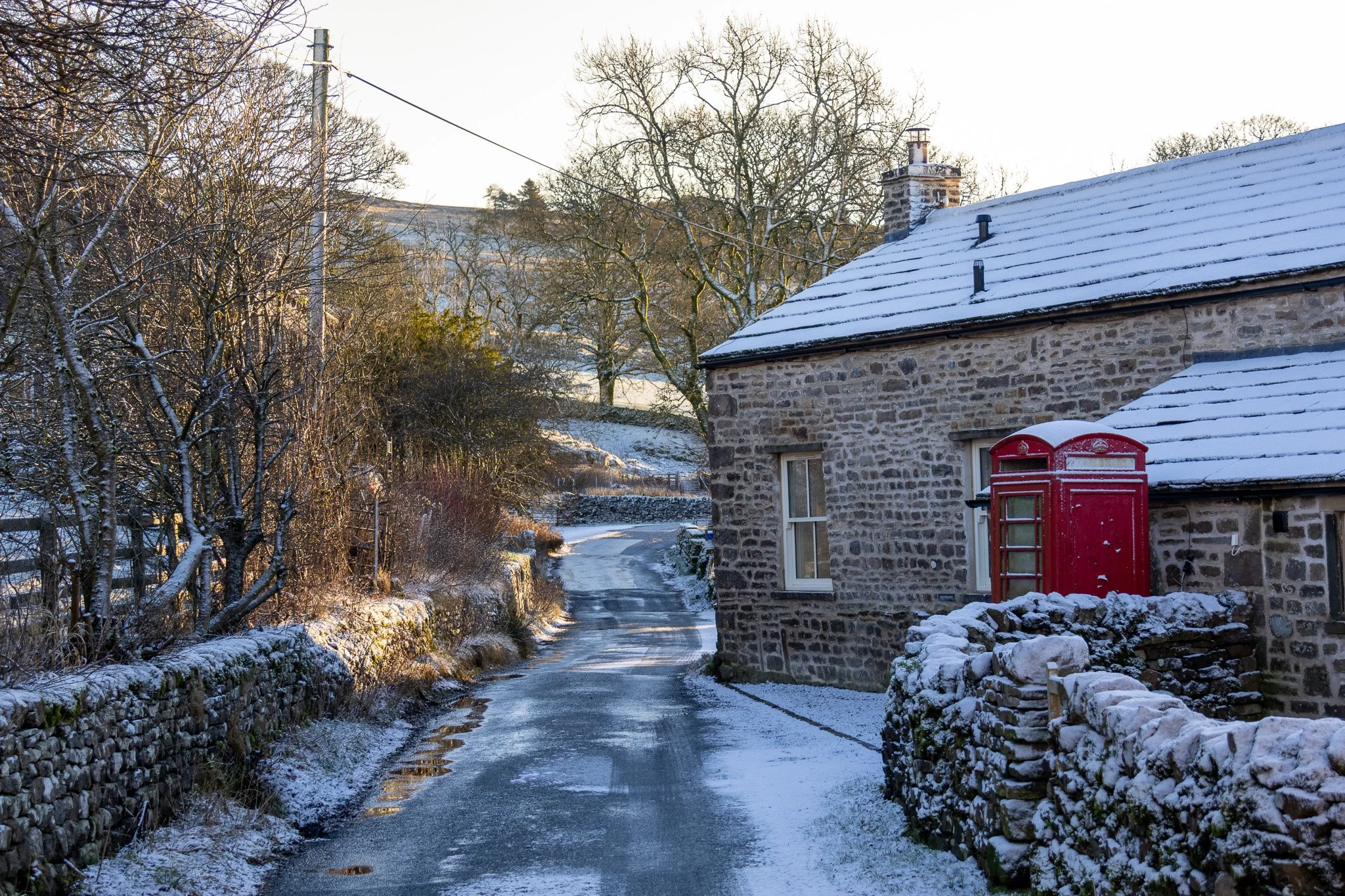 A snow-covered country road adjacent to a stone house with a red telephone booth outside, in a rural winter landscape with trees and hills in the background.