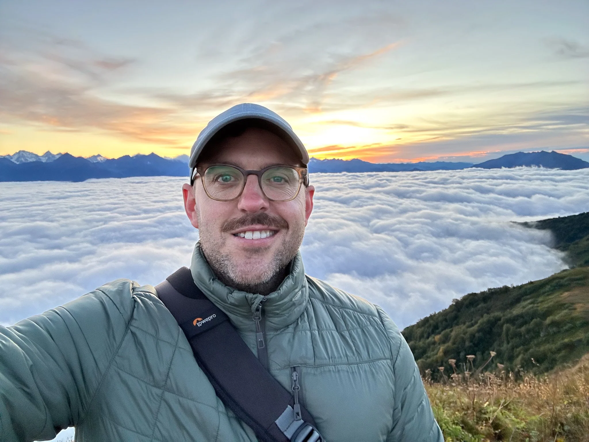 A man in a green jacket, glasses, and a cap taking a selfie at sunrise or sunset on a mountain with clouds below and distant snow-capped peaks.