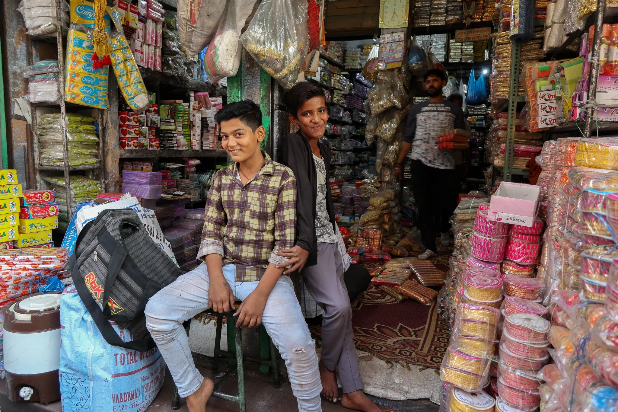 Two boys sitting inside a busy general store with shelves filled with various packaged goods, snacks, and household items, and another person walking in the background.