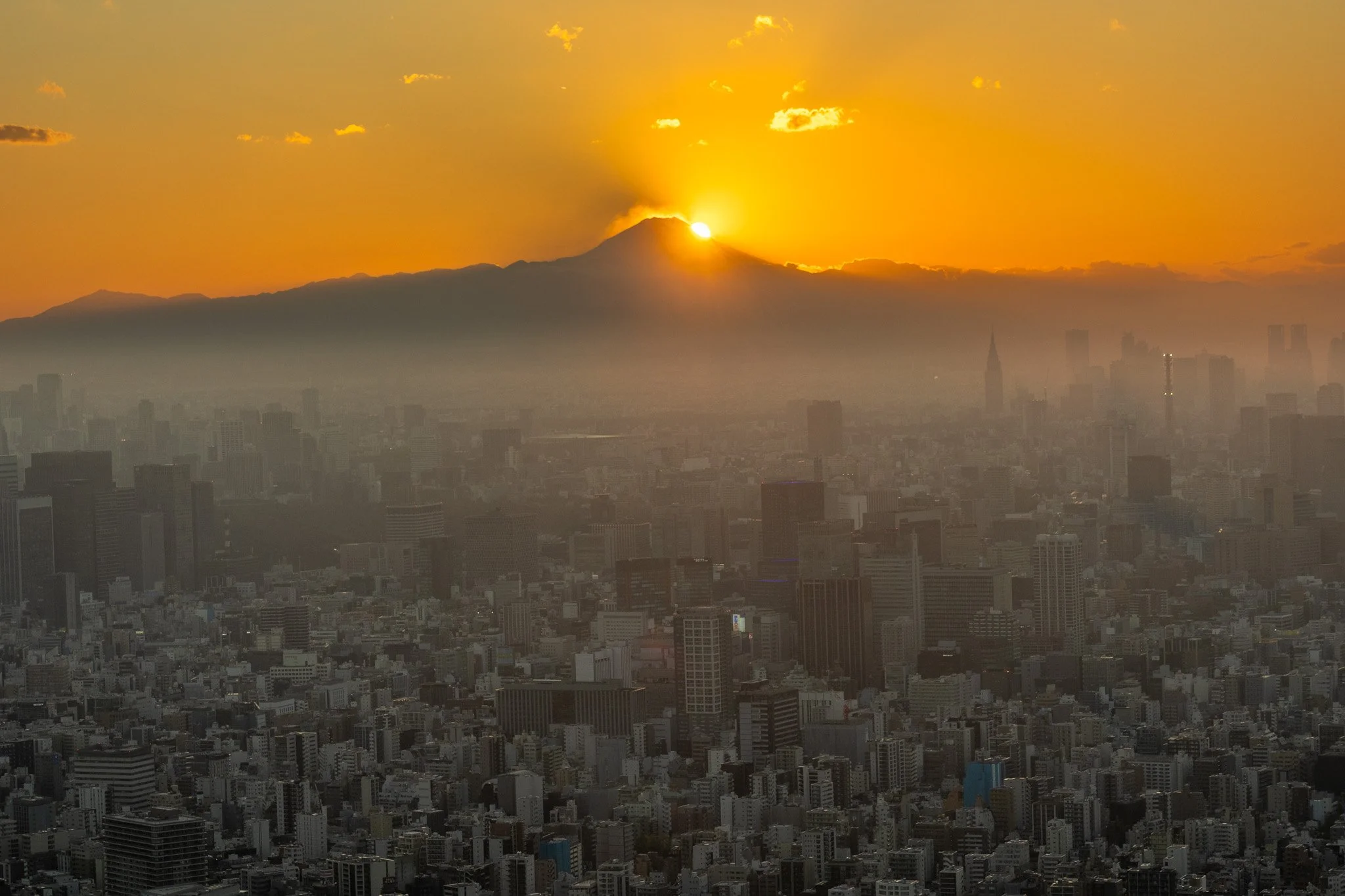 City skyline at sunset with Mount Fuji in the background, partially obscured by clouds and fog.