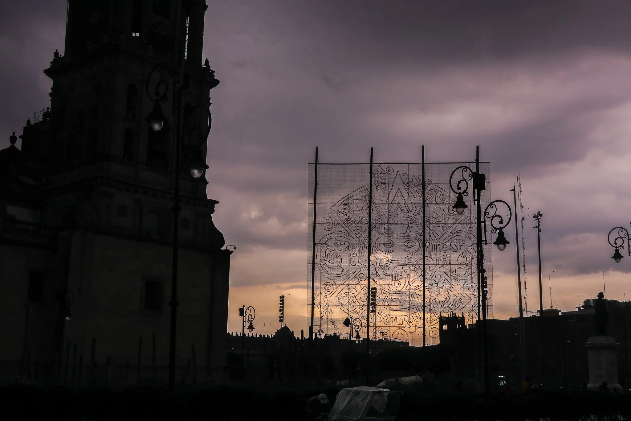 Dark silhouette of a historic building with a tall clock tower, illuminated street lamps, a decorative metal framework, and dark stormy clouds in the sky at dusk.