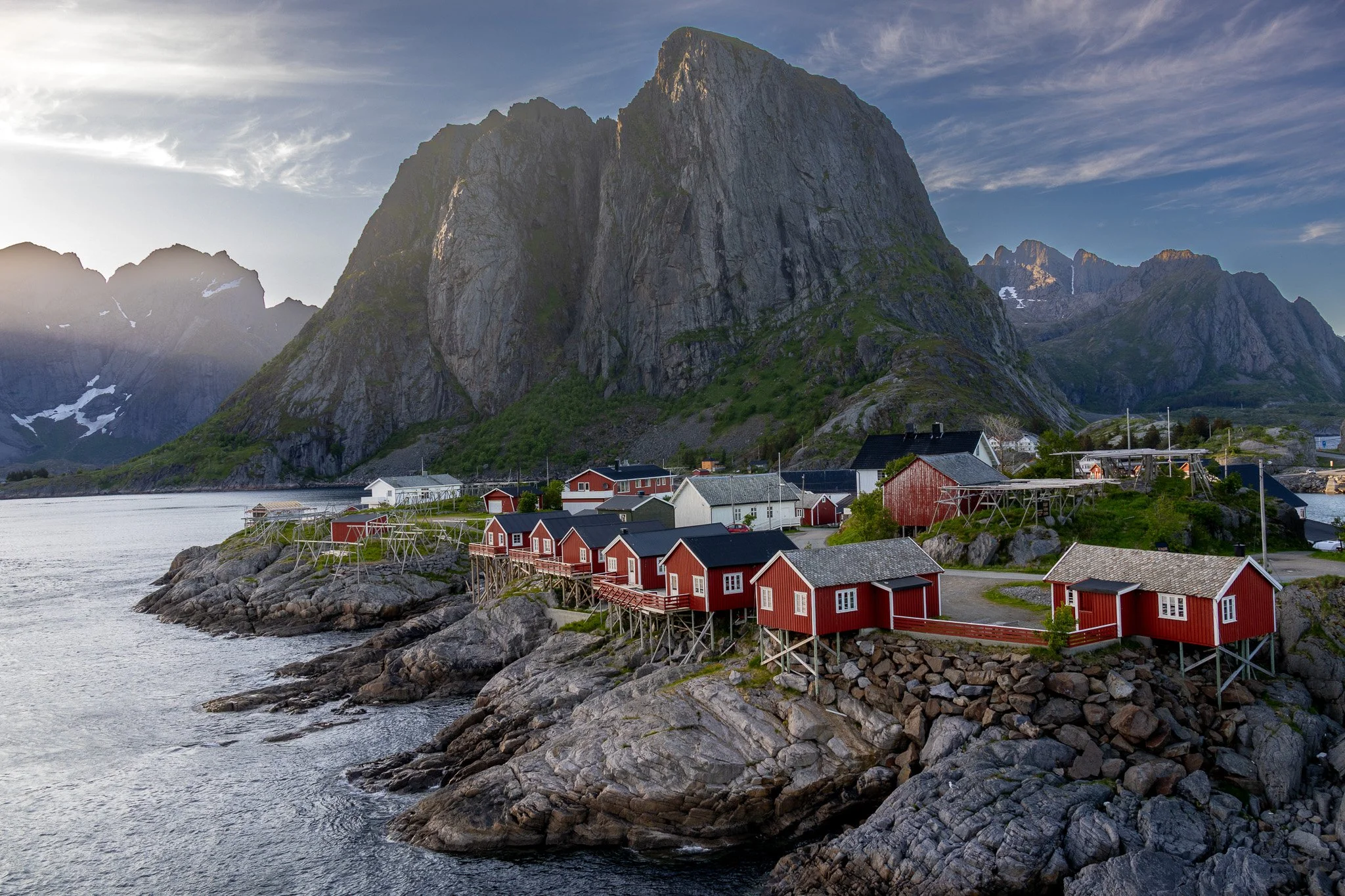 Coastal village with red wooden houses on rocky shore, surrounded by mountains under a partly cloudy sky.