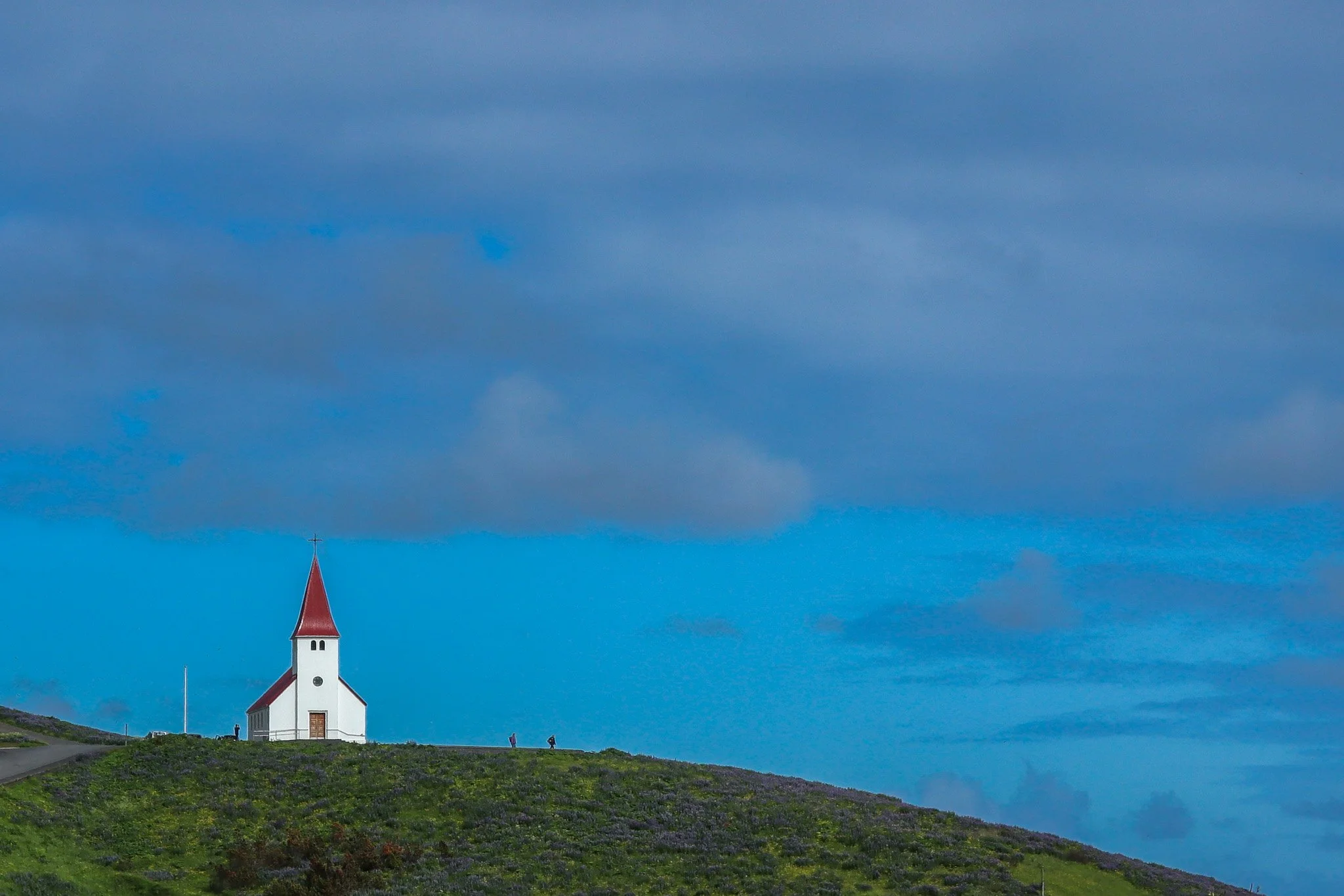 Small white church with red steeple on a grassy hill against a blue sky with gray clouds.