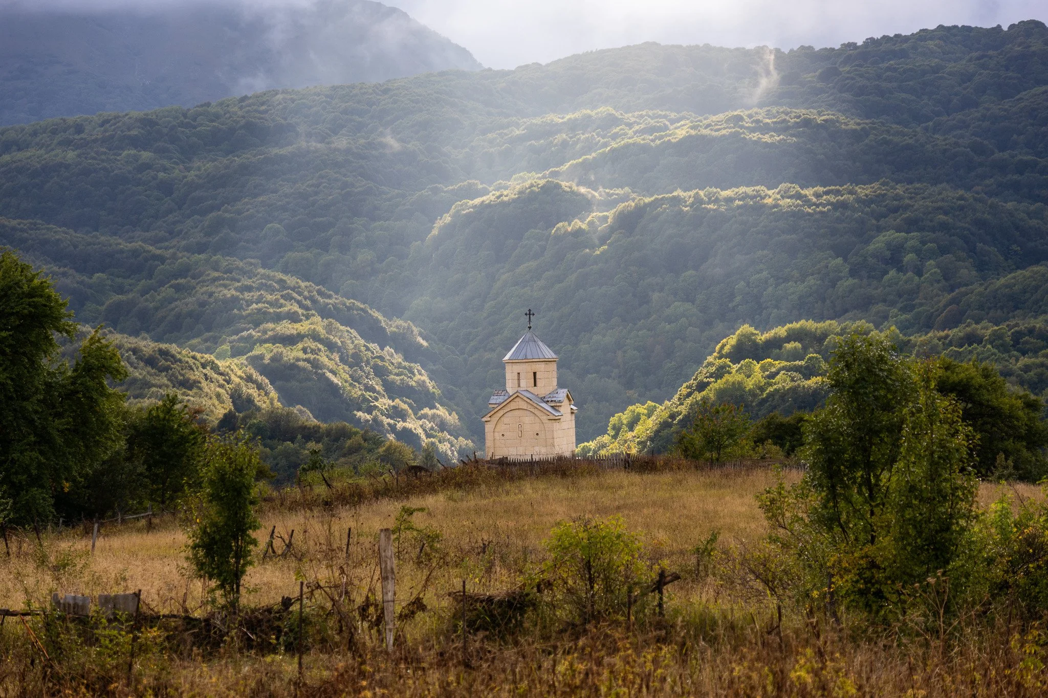 A small, white church with a cross on top, situated in a grassy field with trees, against a backdrop of forested mountains and a partly cloudy sky.