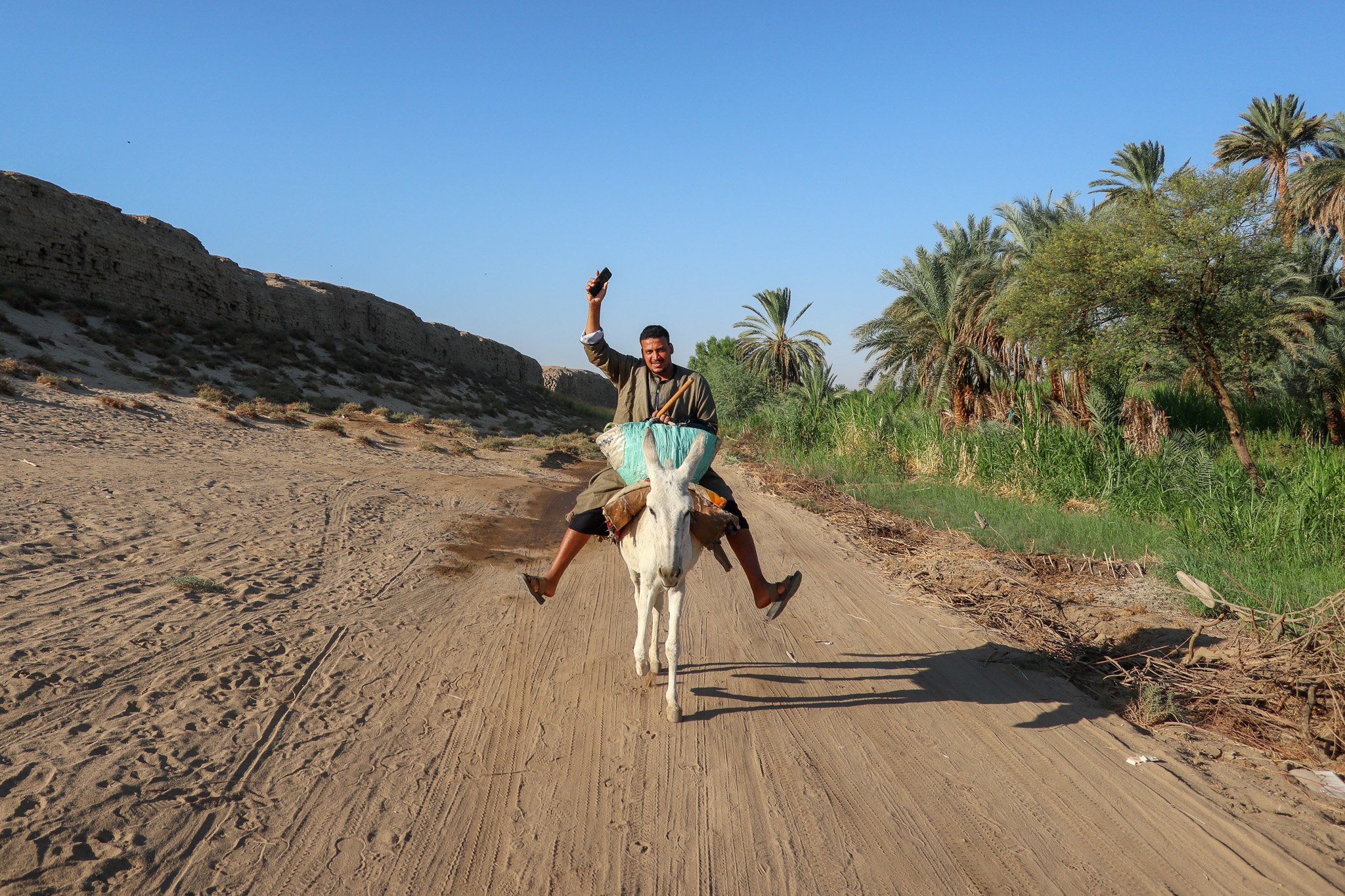 A man riding a donkey on a dirt road, raising his right hand with a smile, in a desert-like area with palm trees and green plants in the background and clear blue sky.