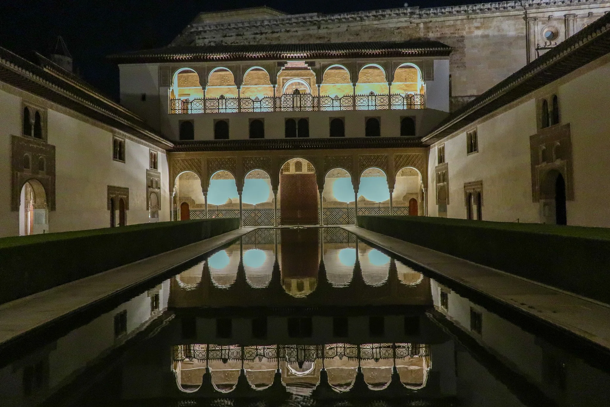 Exterior view of a historic building at night with illuminated arches and balconies, reflecting in still water in the courtyard.