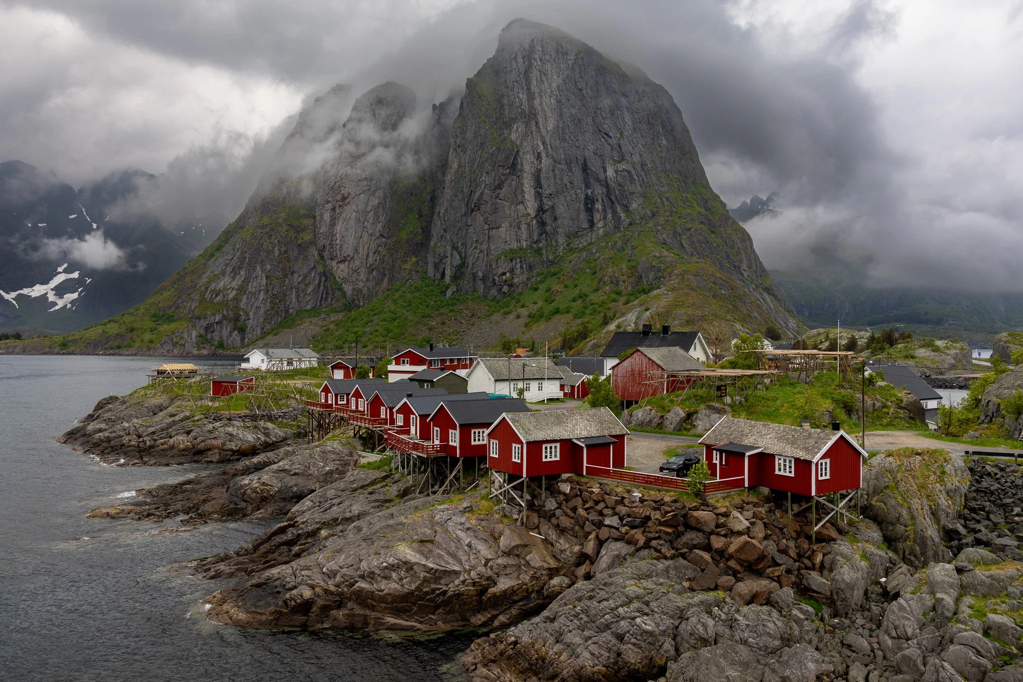 A coastal village with red and white wooden houses on rocky shores, set against a backdrop of large mountains shrouded in clouds.