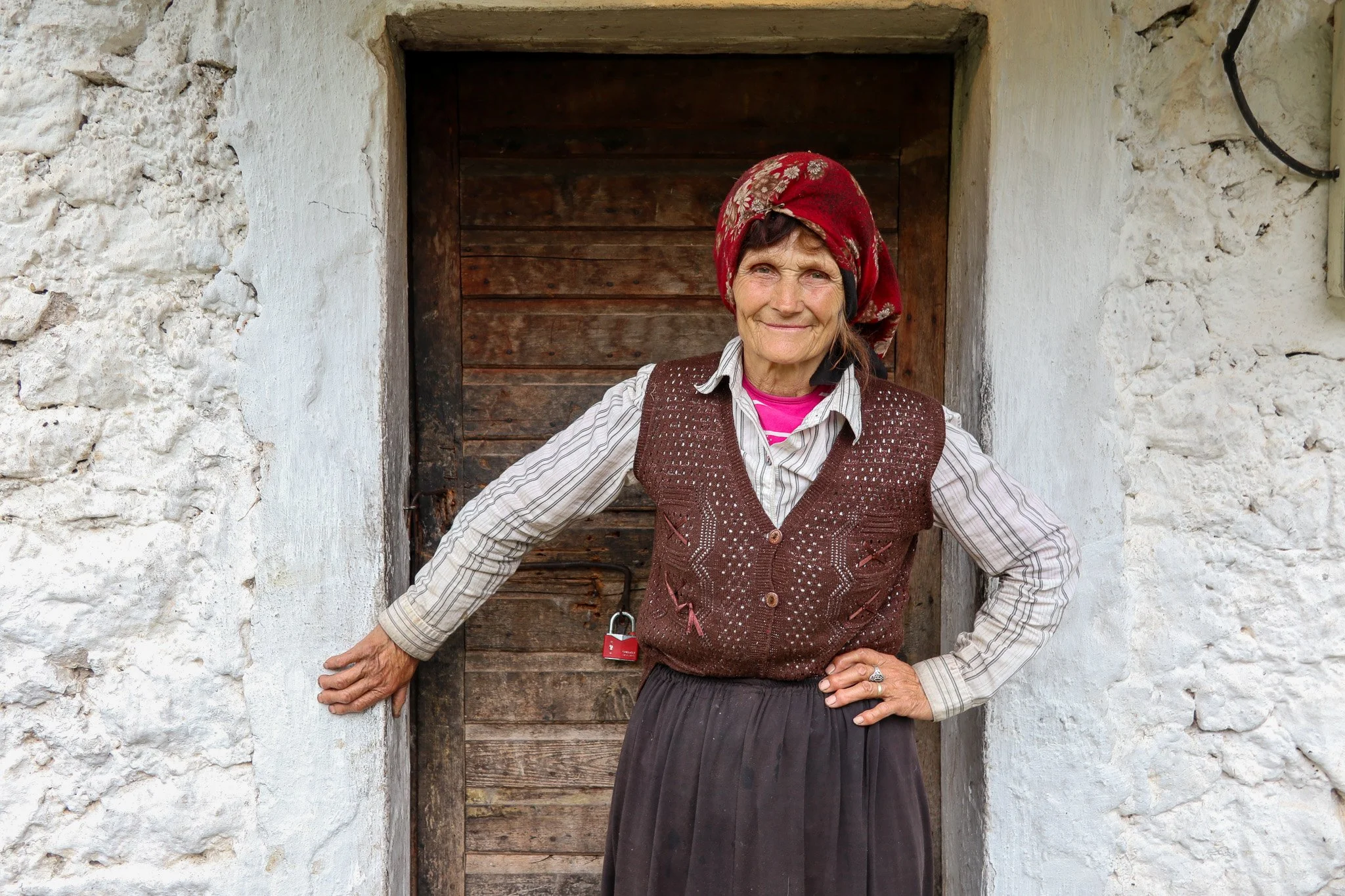 An elderly woman stands in front of a wooden door with white stone walls, wearing a red headscarf, striped shirt, brown vest with patterns, and a long dark skirt, smiling.