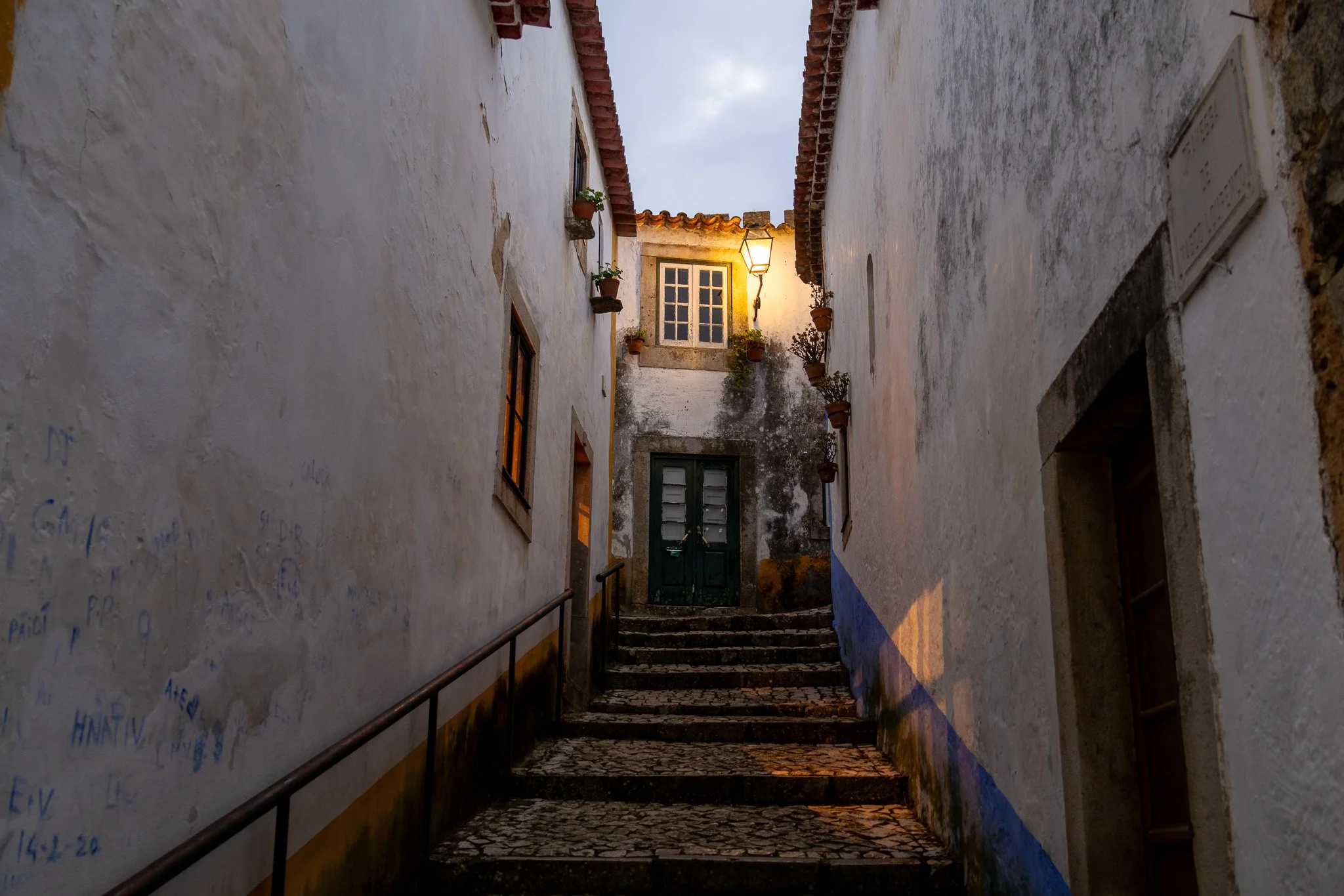 Narrow alleyway with steps leading up, flanked by white stucco buildings with small windows and potted plants, illuminated by a street lamp at dusk.